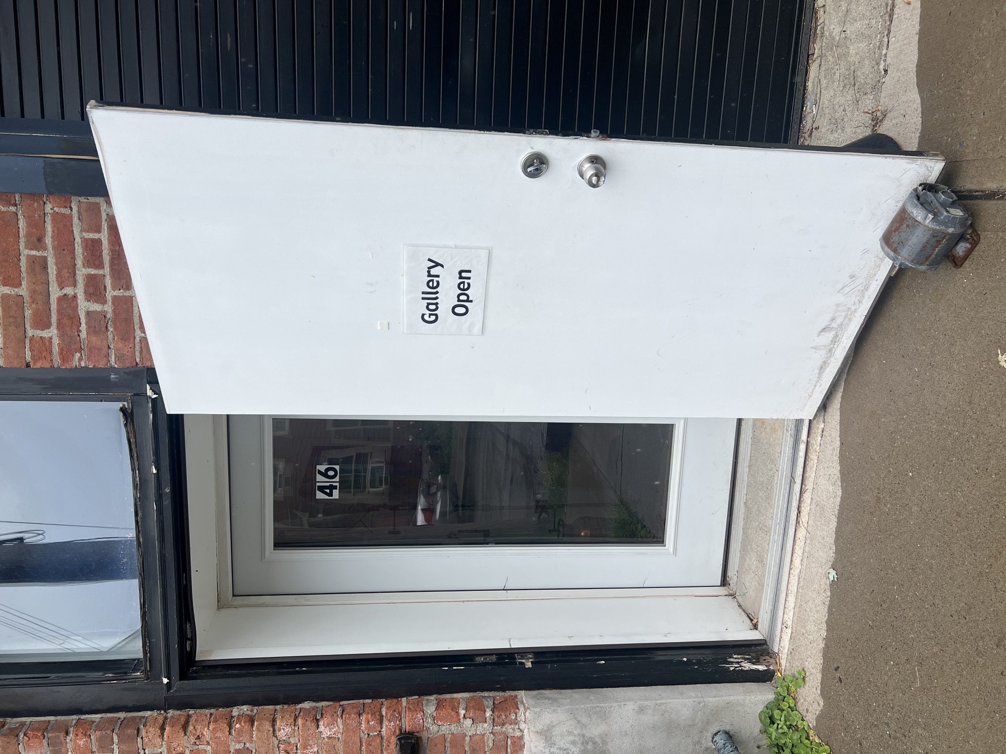 The photograph shows the open door of a business with a sign reading "Gallery Open". The door is white and has two doorknobs. The reflection in the door shows a building across the street with the number 46 on it. The building exterior consists of brick and black trim. There is a rusty metal object on the ground, propping the door open. The scene appears to be in an urban environment, and based on available information, is located in the City of Newburgh, United States of America.