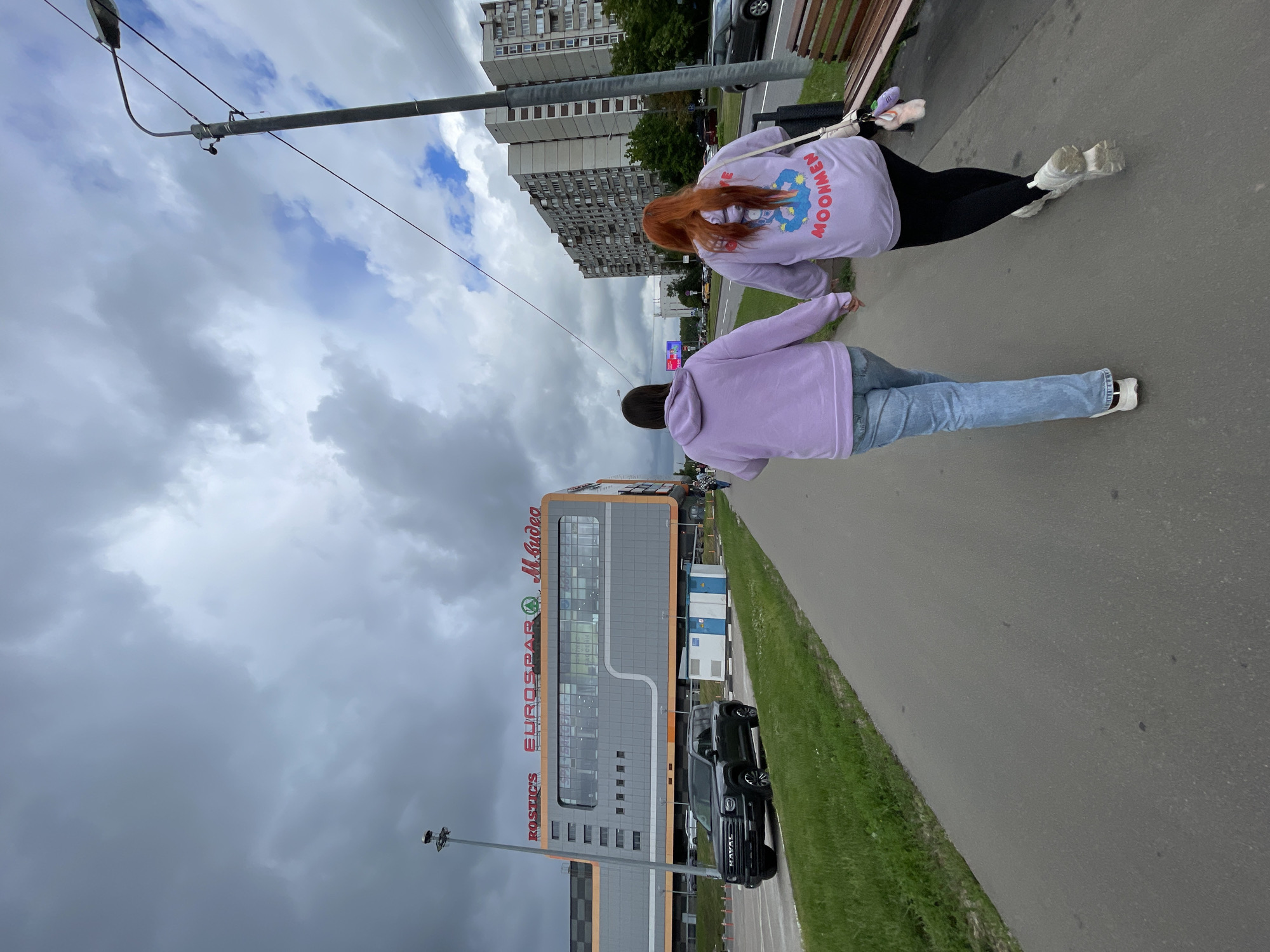 Two individuals, viewed from behind, walk hand-in-hand along a paved pedestrian walkway. They are both wearing casual clothing, with one in a light purple hoodie and blue jeans, and the other in a similar purple hoodie with a graphic design and black leggings. To their left, a grassy area borders a road with a dark-colored Haval SUV parked on it. Beyond the road, a large, modern commercial building with orange and grey facade is visible, bearing signs for "KOSTICS," "EUROSPAR," and "M.Video." In the background, tall residential buildings and a streetlamp dominate the urban landscape under a cloudy sky. The scene is consistent with an outdoor environment in a city, likely in Moscow, Russia, given the architectural style and signage.