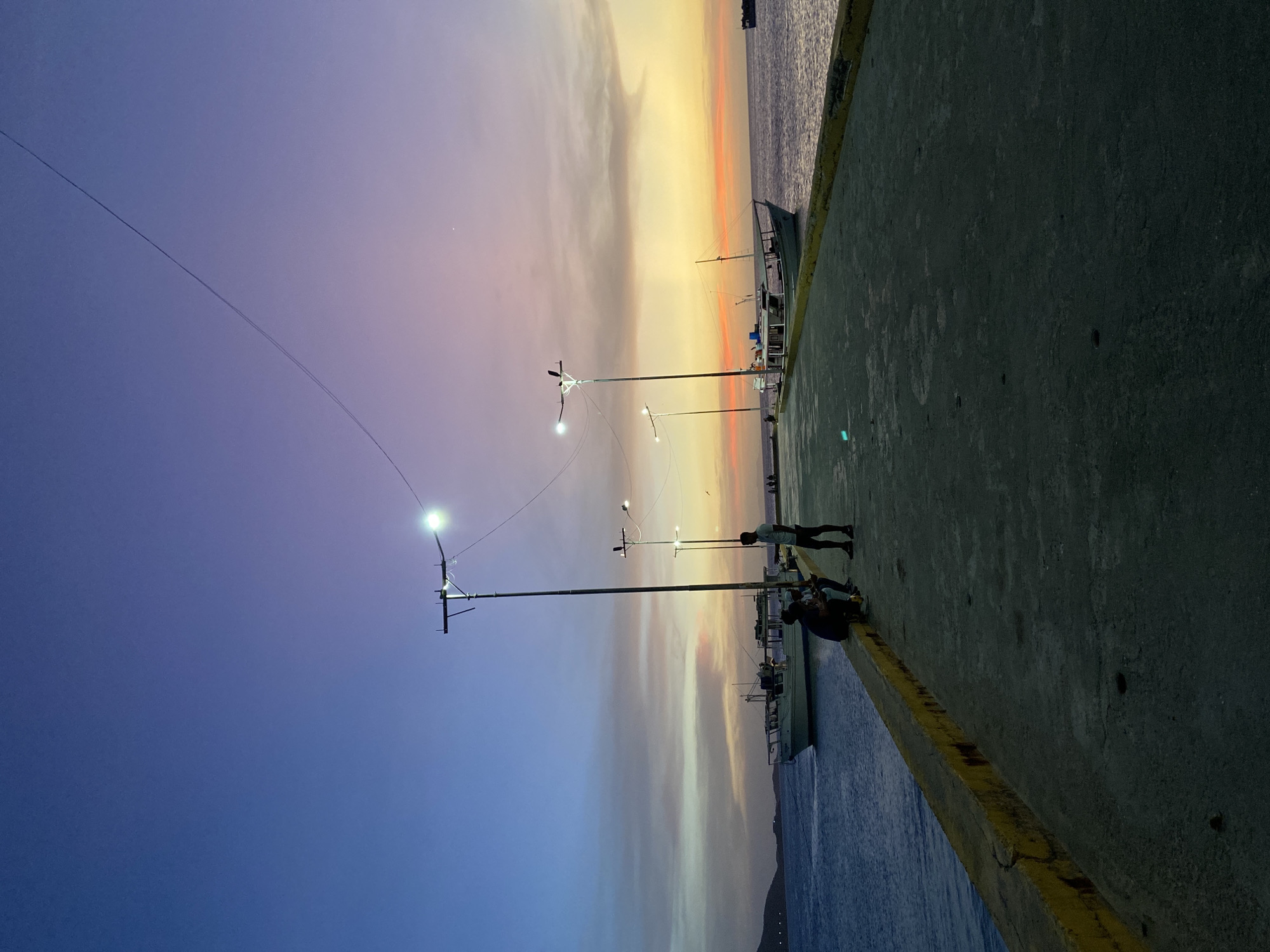 The image depicts a sunset over the water in Juan Griego, Venezuela. Two people are sitting near the edge of a pier with their legs hanging over the side, while another person is standing nearby. Fishing boats are docked along the pier. Streetlights illuminate the pier. The sky is a gradient of colors, from purple and blue to orange and yellow.