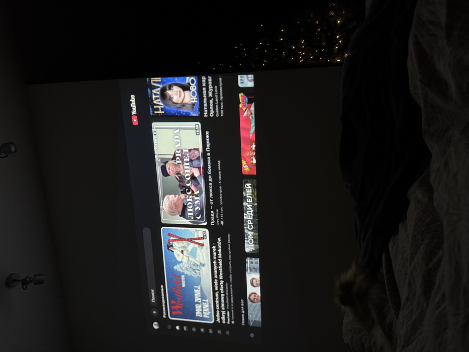The image shows a YouTube screen on a TV. The YouTube interface is in Russian, displaying video recommendations. Some of the recommended videos are "Natalia" a channel of Orlov, Zhuravl; "Prada - from luxury to bum in Paris" by Grey Wiese; and "One center, many famous brands - discover the winter offer of Westfield Mokotów." The search field is visible at the bottom of the screen, and the YouTube logo is in the top left corner.