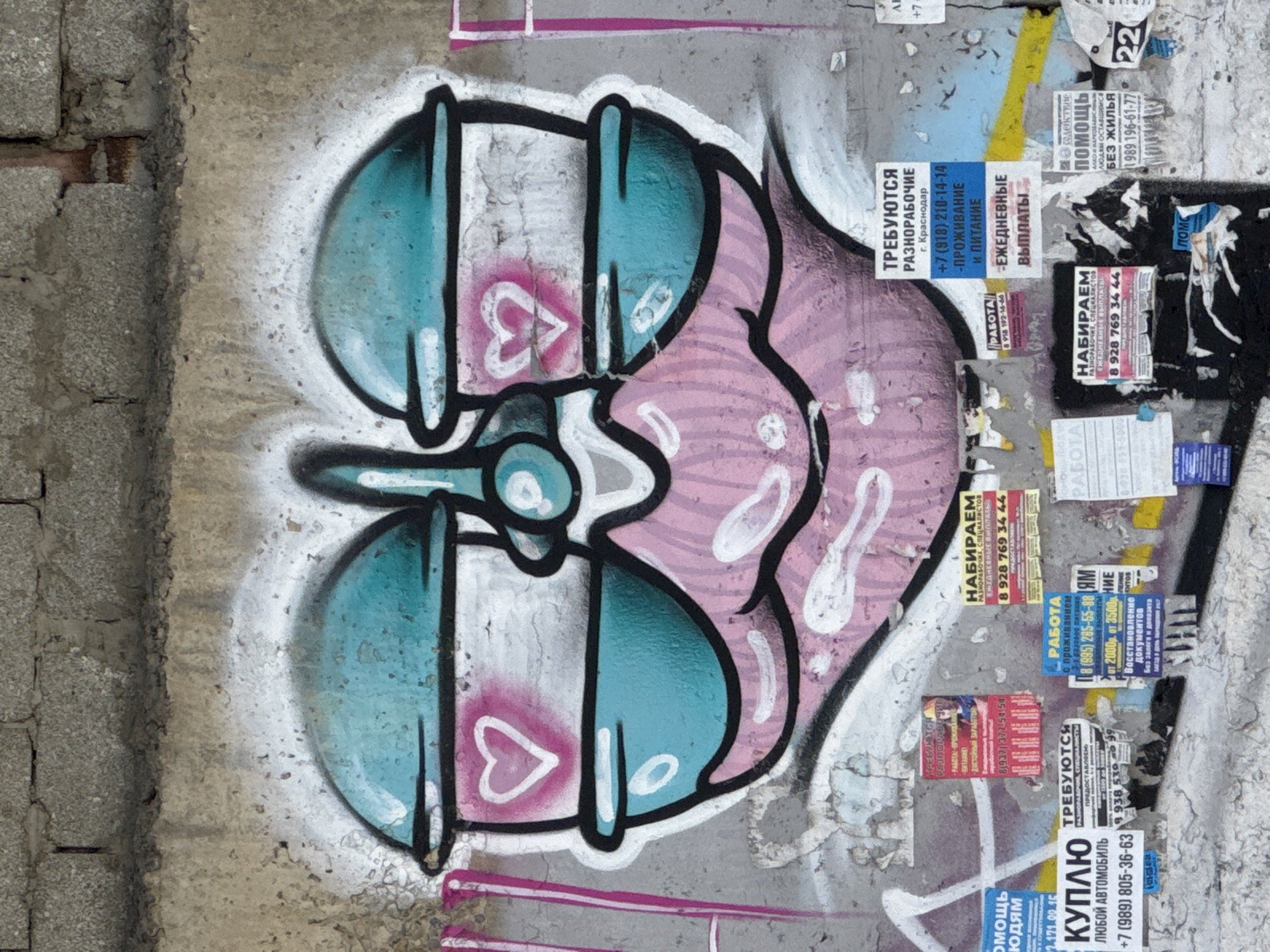 The image shows a close-up of a weathered wall in Krasnodar, Russia, covered in graffiti and assorted advertisements and notices. The graffiti depicts a stylized, symmetrical design in shades of blue and pink, outlined in black, with heart motifs incorporated. Below the graffiti, numerous small posters and leaflets are pasted onto the wall, advertising job opportunities ("ТРЕБУЮТСЯ" - "Required"), housing assistance ("ПОМОЩЬ ЛЮДЯМ" - "Help to People"), and other services. Phone numbers are prominently displayed on the notices. The wall itself appears to be made of concrete blocks and is stained with age and weather.