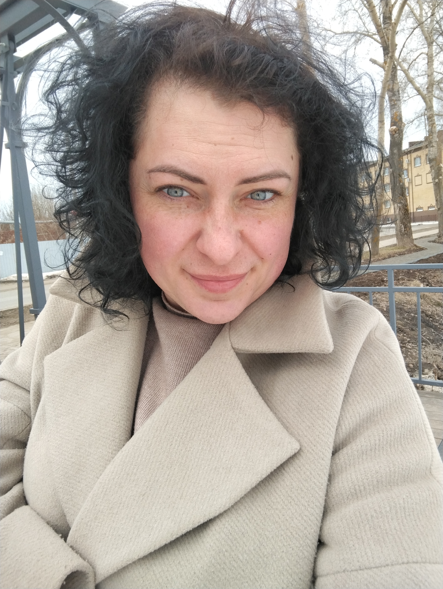 A selfie photograph shows a woman with dark, curly hair and blue eyes, wearing a tan coat and turtleneck sweater. She is outside, likely in Votkinsk, Russia, as evidenced by the architecture of the buildings and landscape in the background. Behind her is a metal structure, possibly part of a pergola or bus stop. Bare trees are visible in the background, along with a sidewalk and a building with light-colored walls and dark windows. The weather appears to be overcast or cold.