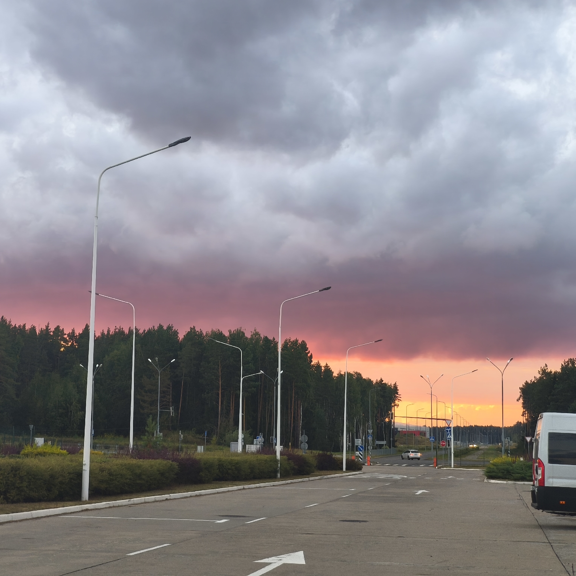 The image depicts an outdoor scene at dusk, likely near an airport or industrial area, as indicated by the presence of numerous tall streetlights lining a paved roadway. A dense forest of green trees forms a backdrop across the majority of the image. The sky above is dramatic, characterized by heavy, dark gray clouds that are dramatically illuminated from below by a vibrant, reddish-pink and orange sunset. In the foreground, the asphalt road is marked with white directional arrows. To the right, a portion of a white van is visible. In the distance, several white cars and more streetlights are discernible. The location is identified as Драчкаўскі сельскі Савет, Belarus.