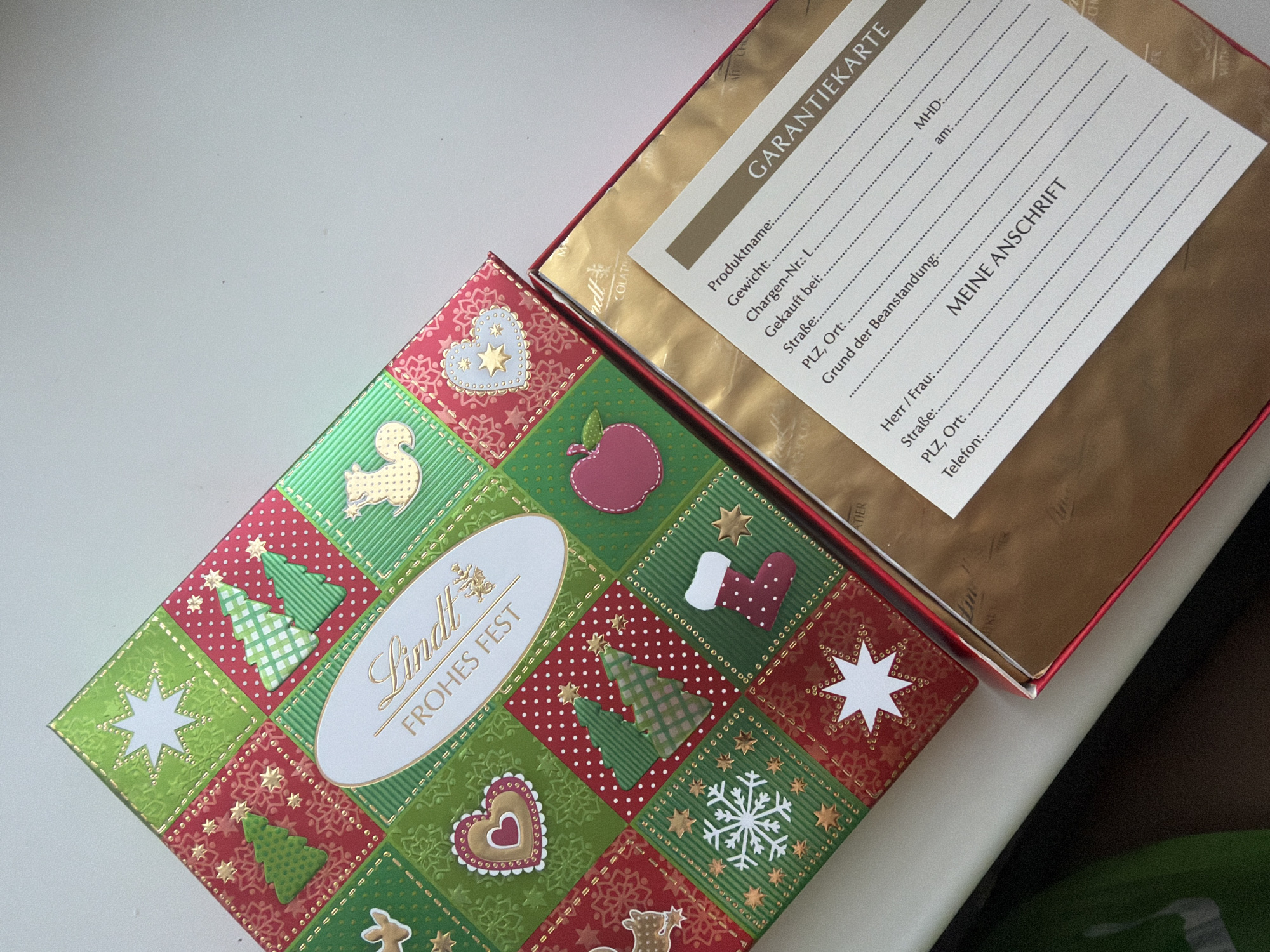An open, rectangular Lindt chocolate box is positioned diagonally on a light-colored surface in Perm, Russia. The box lid features a festive, patchwork design with alternating red and green squares, each containing a Christmas-themed motif such as trees, stars, hearts, a snowflake, a stocking, a reindeer, a squirrel, and an apple, all embellished with gold accents. A central white oval label prominently displays the "Lindt" logo and the German phrase "FROHES FEST" (Merry Christmas). The interior of the box is lined with reflective gold metallic paper. A white "GARANTIEKARTE" (Warranty Card) is visible inside the box, listing fields in German for "Produktname" (Product name), "Gewicht" (Weight), "Chargen-Nr." (Batch No.), "Gekauft bei" (Purchased from), "Straße" (Street), "PLZ, Ort" (Postal code, City), "Grund der Beanstandung" (Reason for complaint), "MHD" (Best before date), "am" (on date), and "MEINE ANSCHRIFT" (My Address) with corresponding fields for personal details. No individuals are present in the scene.