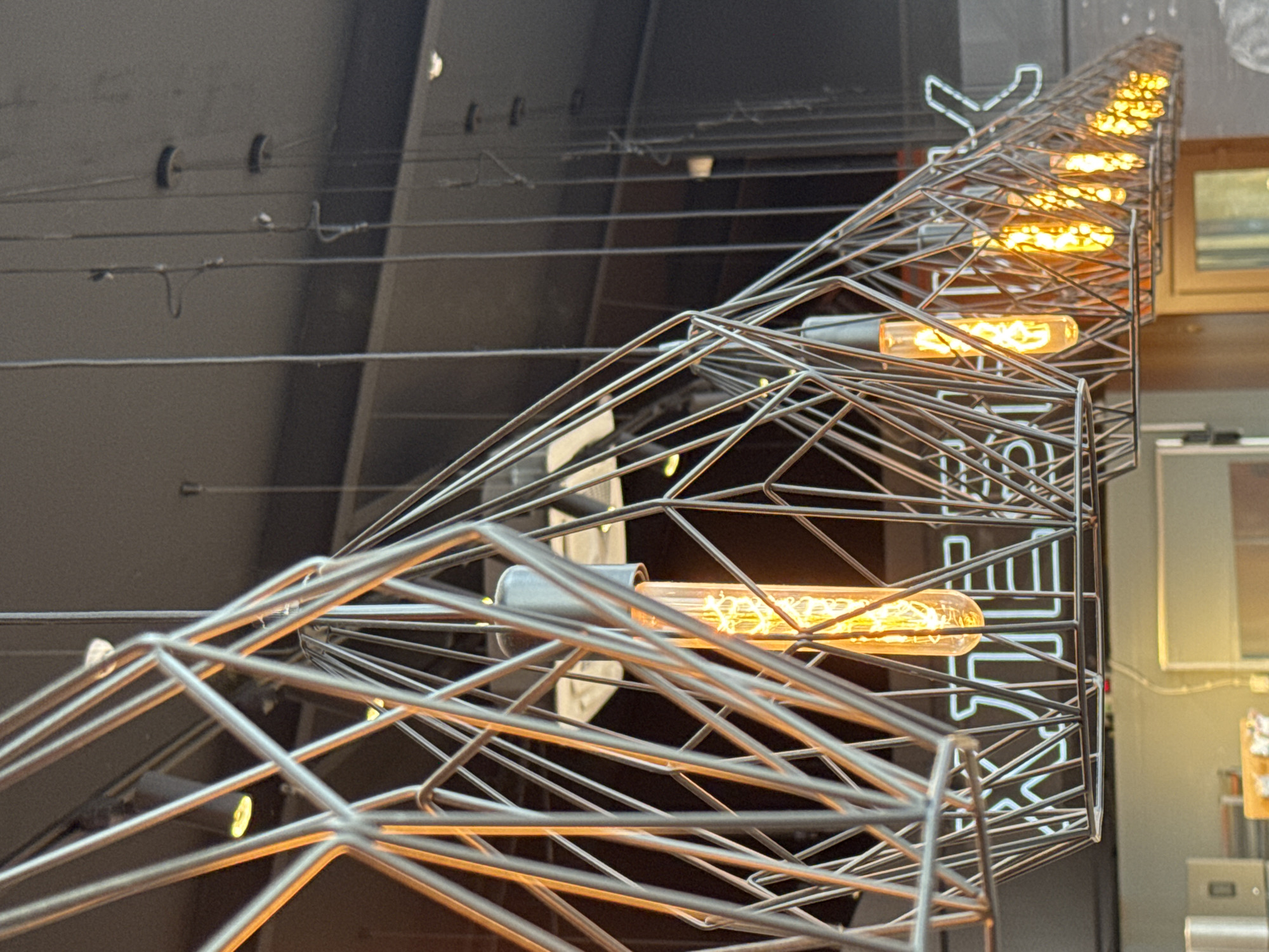 The image shows a decorative lighting fixture composed of a metallic, geometric frame housing a series of elongated, warm-toned light bulbs. The framework is intricate, with numerous intersecting wires creating a cage-like appearance. The bulbs emit a soft, orange light, providing a stark contrast to the cool, metallic tones of the structure. The background features indistinct architectural elements and a partially visible sign, suggestive of an indoor commercial space. While the prompt mentions the city of Saint Petersburg, Russia, there are no discernible features within the image that specifically confirm this location.