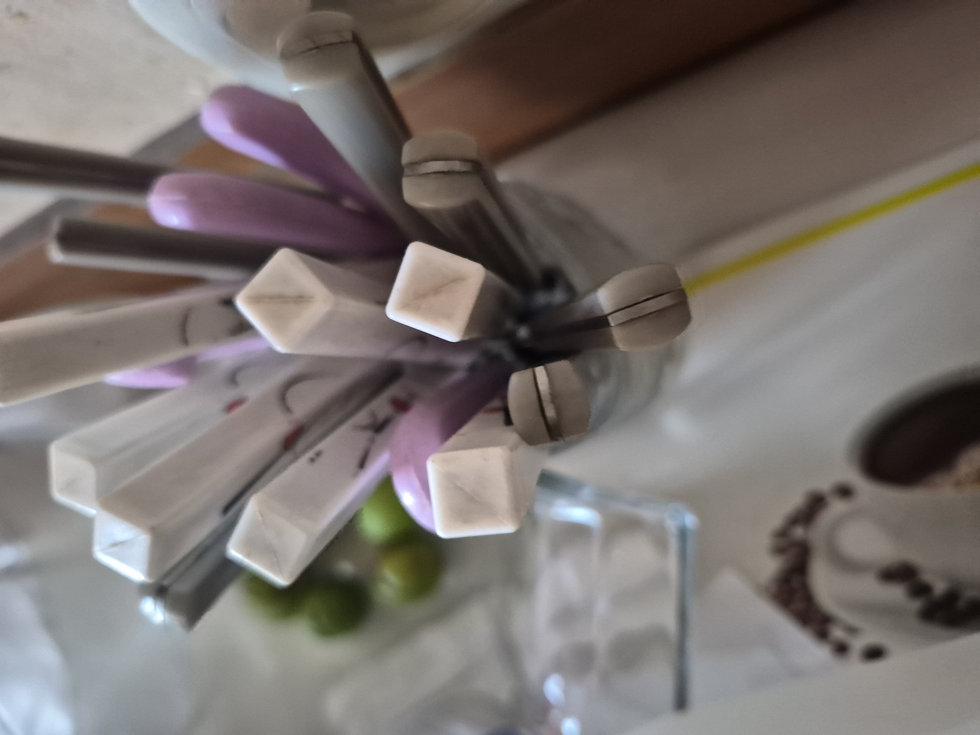 The image provides an overhead view of a collection of chopsticks held upright in a clear glass container on a light-colored surface. The chopsticks vary in color, featuring shades of white, grey, light purple, and pink. Many of the grey chopsticks have distinct metallic rings near their rounded ends, while several white ones exhibit square ends. Upon closer inspection, some white chopsticks have faint decorative elements, including subtle black characters or symbols, and one appears to show part of a red heart design.
In the foreground, below the chopstick holder, several green limes are visible, slightly out of focus. To the right side of the frame, a white cup containing a dark liquid, presumably coffee, is accompanied by scattered brown coffee beans, both rendered with a soft focus. A bright yellow linear object, possibly a straw or a decorative strip, runs diagonally in the blurred background. The scene suggests an indoor setting, likely a kitchen or dining area, with items indicative of a meal or beverage service. The activity implied is either preparation for or the aftermath of dining. While the provided context specifies the location as Yerevan, Armenia, there are no visual cues within the image to confirm this.