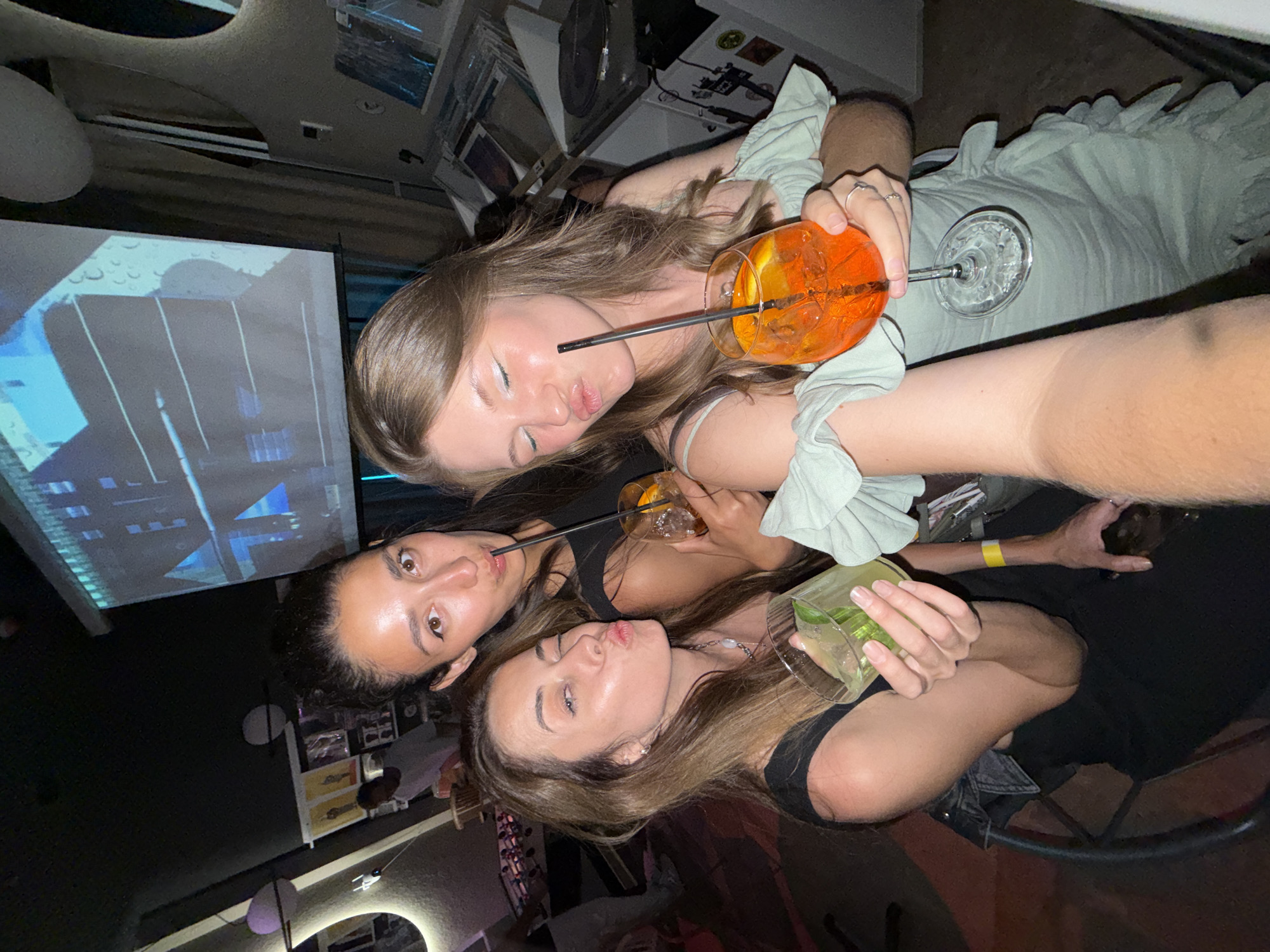 The image depicts three women in a dimly lit indoor bar or club setting, located in Rafailovići, Montenegro. The woman on the viewer's left has medium brown hair, wears a dark top, and holds a clear glass containing a green cocktail with mint leaves and lime; she poses with pursed lips and a partially closed right eye. The central woman has dark hair pulled back, wears a black top, and holds a glass with an amber-colored drink, an orange slice, and a black straw; she also poses with pursed lips and wears a yellow wristband on her right arm. The woman on the viewer's right is taking a selfie, extending her arm forward; she has light brown hair, wears a light green off-the-shoulder dress with ruffled details, and holds a stemmed glass containing an orange Aperol Spritz with ice, an orange slice, and a black straw, with her eyes closed and lips pursed. In the background, a large projection screen displays blue and grey geometric patterns. Textured, light-colored wall structures are visible on both sides of the frame. A male figure in a white shirt is partially visible behind the women to the left. To the right, shelves holding media, likely records, and a turntable are present.