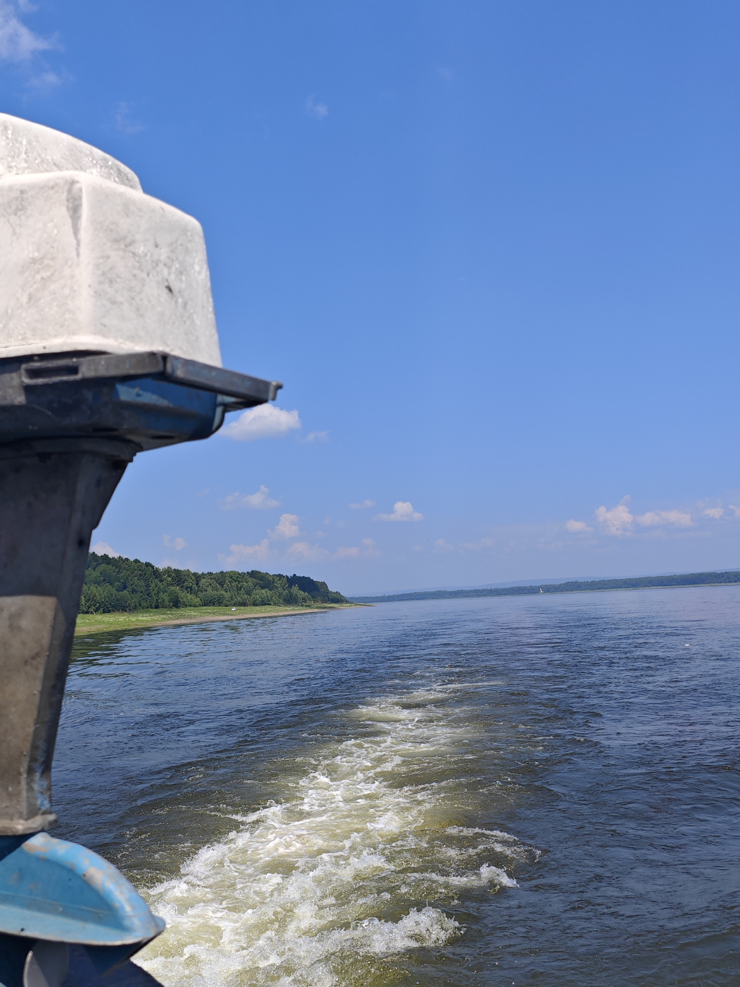An outboard motor of a boat, partially visible in the foreground on the left, propels the vessel through a large body of water, leaving a white wake. The water is a deep blue, with the wake appearing foamy and disturbed. In the distance, a tree-lined shore stretches horizontally across the frame, with a narrow strip of lighter green land visible along the water's edge. The sky above is a vibrant, clear blue with a few scattered white clouds. The overall impression is that of a sunny day on the water, likely on a river or lake, with the boat moving away from the viewer and towards the distant shore. No people are visible in the image. The location is the Ob River, near the village of Pogodaevo, in the Novosibirsk Oblast of Russia.