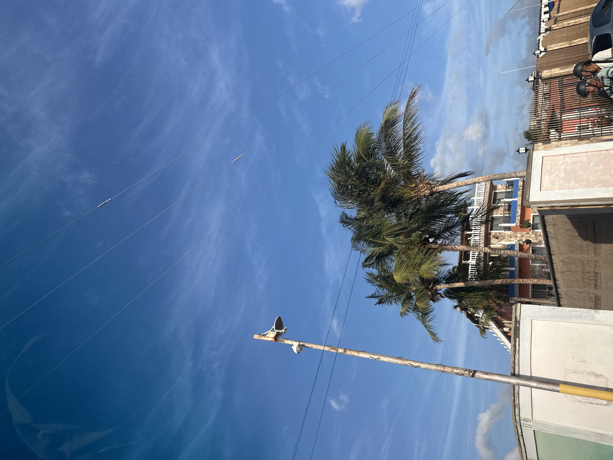In Juan Griego, Venezuela, a clear blue sky with wispy white clouds forms the backdrop for a street scene. In the foreground, a rustic wooden pole stands tall, supporting a security camera and what appears to be a satellite dish. Several power lines are strung across the sky, running diagonally from the upper left to the lower right. Behind the pole, a multi-story building with a colorful facade, featuring stone accents and balconies, is visible. Several palm trees with lush green fronds are scattered around the building, their leaves rustling in the breeze. A low wall separates the street from the property. Further to the right, a decorative metal fence with bamboo-like accents encloses the property. Two individuals wearing helmets are seated on a motorcycle in the lower right corner of the frame, suggesting an outdoor urban setting.