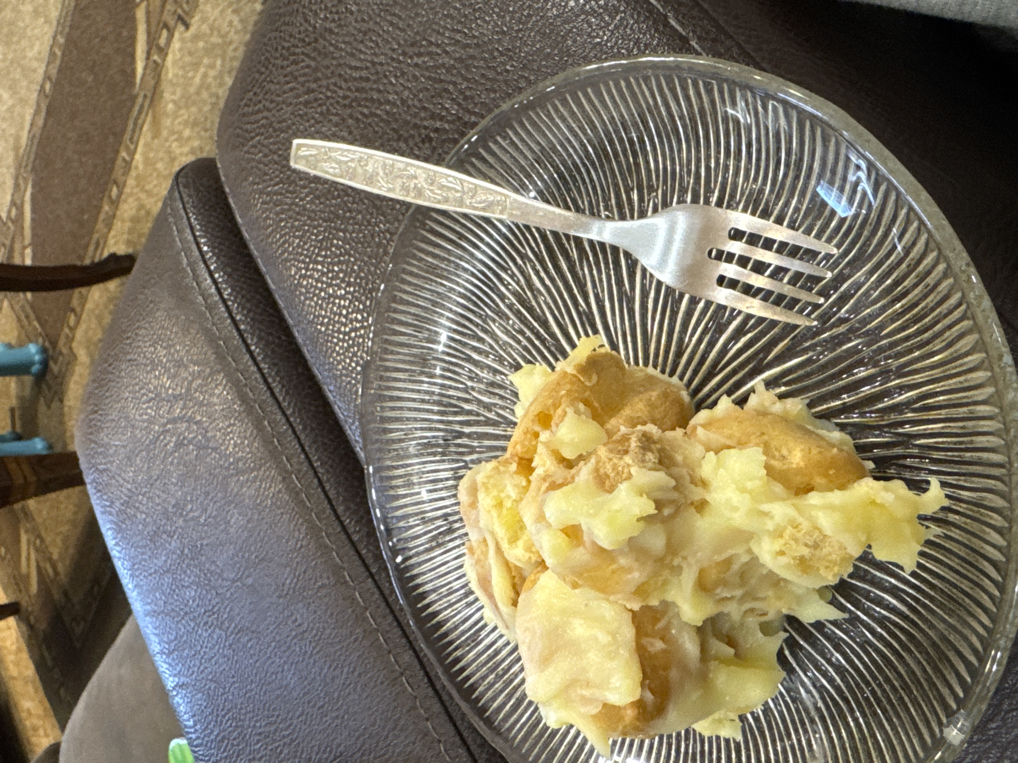A dessert is in a clear plate, with a metal fork on top, resting on a dark brown leather or imitation leather cushion. The dessert appears to be a pile of pastry with a creamy or sugary glaze. The clear plate has a spiral groove design. The cushion is part of a chair. It cannot be determined whether the location is in Yerevan, Armenia.