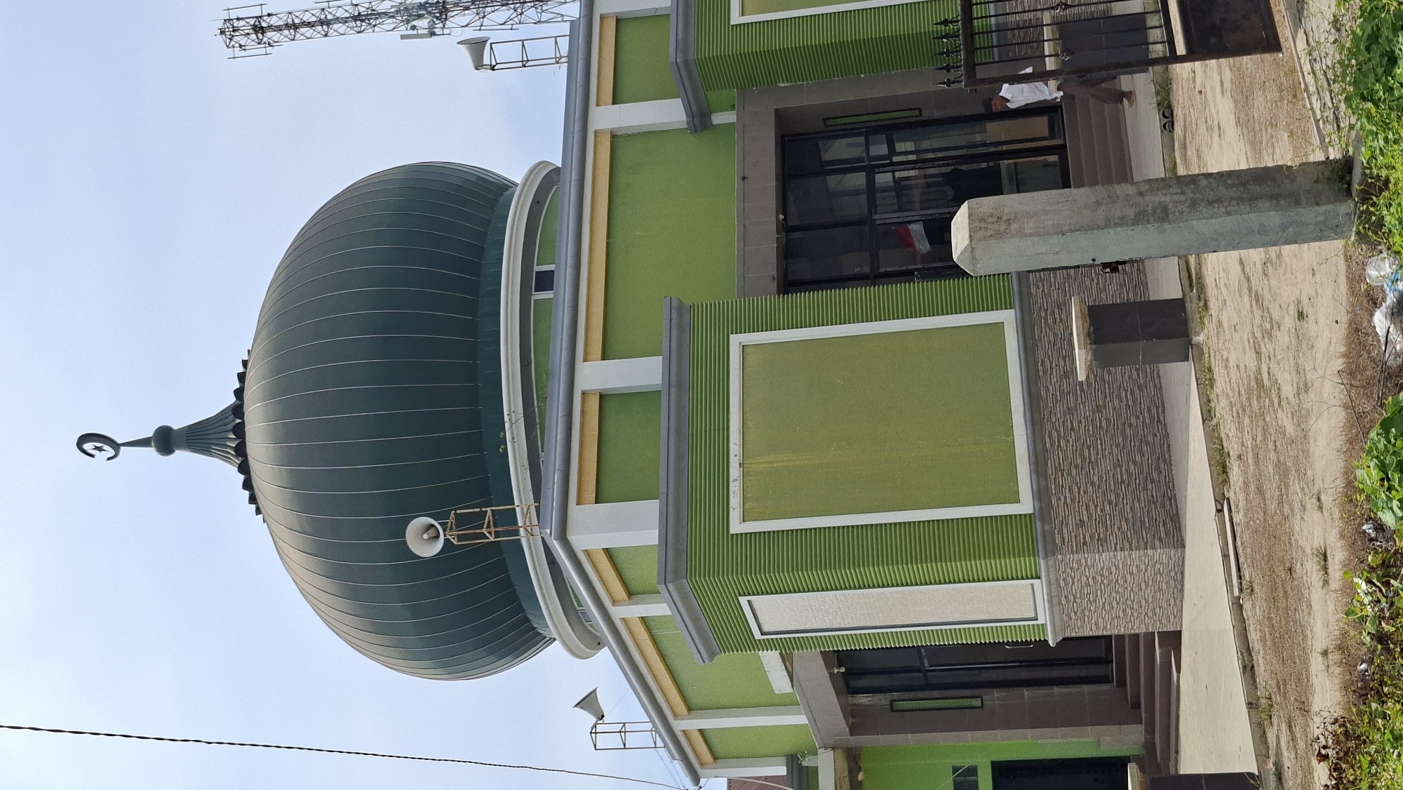 The image shows a mosque in Keudegeudong, Indonesia. The mosque features a large green dome topped with a crescent moon symbol. White speakers are mounted on either side of the dome. The lower part of the mosque is painted green with textured panels and decorative stone-like accents at the base. A person in a white shirt and brown pants is walking near the entrance, which has glass doors. A cell tower is visible in the background to the right. The foreground shows a dirt path, some sparse vegetation, and a concrete pillar.