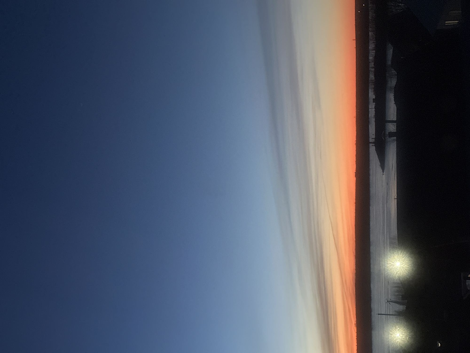 The image shows a sunset in Chaikovsky, Russia. The sky transitions from dark blue to orange near the horizon, with clouds forming vertical bands. A dark landmass with trees and buildings runs along the right side, including two visible lights.