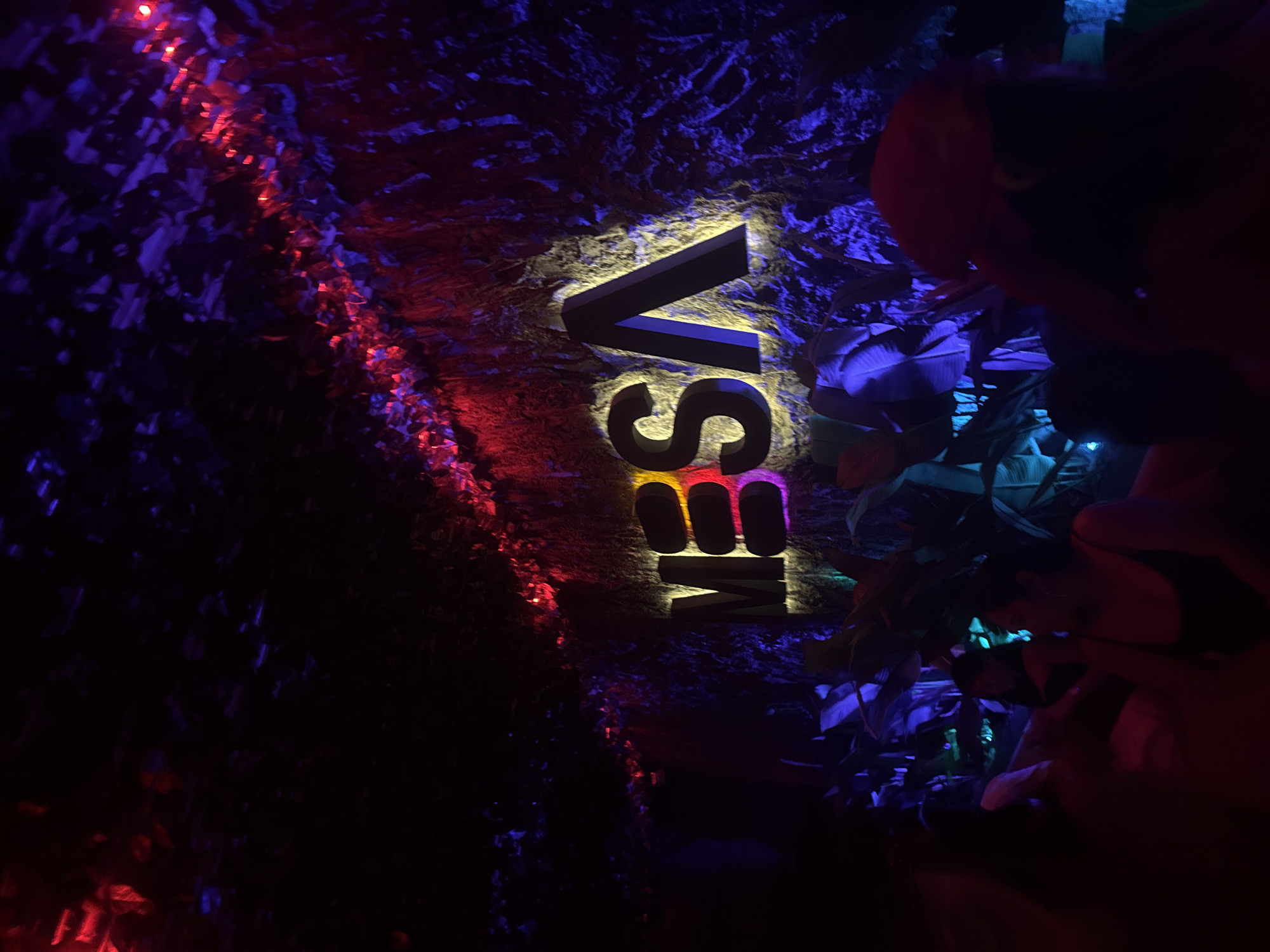 The image captures a dimly lit interior, likely a bar or club, with neon lights illuminating the word "MESA" in large, stylized letters on a textured wall. The wall appears to be made of a material resembling rough stone or bark, and it is adorned with red and purple lighting that creates a dramatic effect. In the foreground, several people are visible, their faces obscured by the low light and their postures suggesting they are seated or standing in a social setting. The atmosphere is dark and vibrant, characteristic of a nightlife venue. This scene is consistent with establishments found in the tourist areas of North Kuta, Indonesia, known for their lively entertainment options.