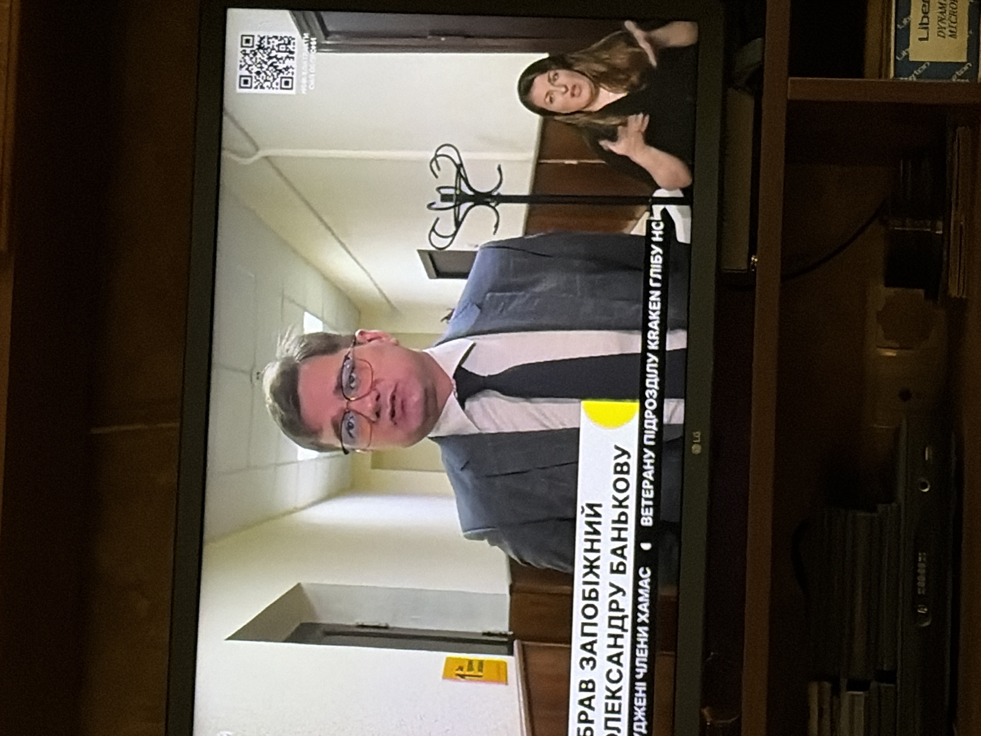 The image shows a television screen displaying a news broadcast. On the left side of the screen, a man wearing glasses and a suit is visible. A caption bar below him reads "БРАВ ЗАПОБІЖНИЙ ОЛЕКСАНДРУ БАНЬКОВУ", with additional text below that is partially obscured. To the right of the man, a woman is gesturing with her hands, appearing to be a sign language interpreter. A QR code is displayed in the top right corner of the screen. The background appears to be an indoor hallway with light-colored walls and doors. The television is placed on a wooden shelf, with various items including what appears to be a DVD player and stacked media visible on the shelf below.