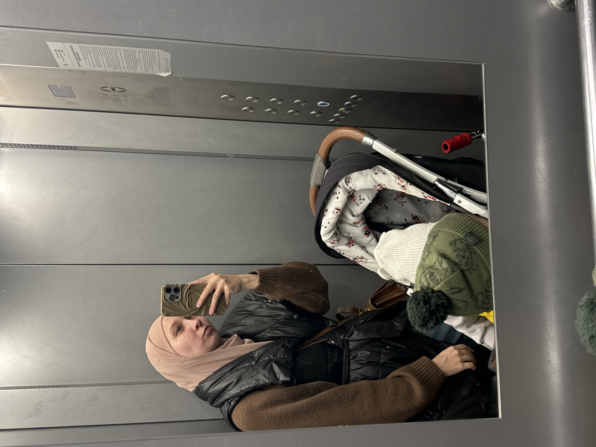 The image depicts a woman taking a selfie in an elevator. She is wearing a headscarf, a brown sweater, and a black puffy jacket. Beside her in the elevator is a stroller filled with clothes and a hat. The elevator has silver walls and a panel of buttons. A sign is visible on the wall above the buttons. The location is the city of Chaikovsky, Russia.