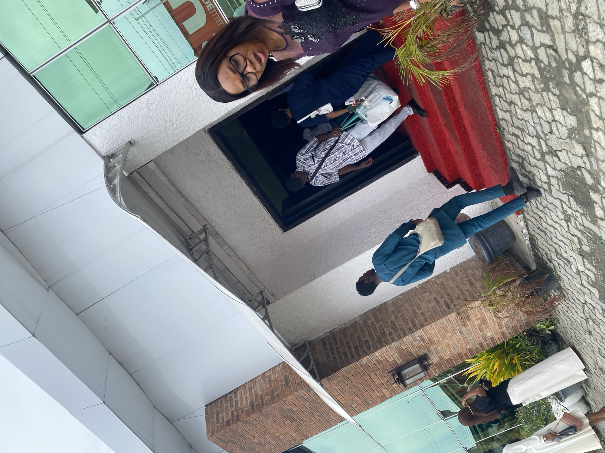 The image depicts an exterior scene likely in Ado, Nigeria. The perspective is a low-angle shot looking upwards at a building facade. People are visible, including one woman wearing glasses and a purple top, positioned near a doorway. Other people are entering the doorway. A man in a blue suit is walking along a paved area. The architecture includes brick and glass elements. Potted plants and decorative elements are present in the area.