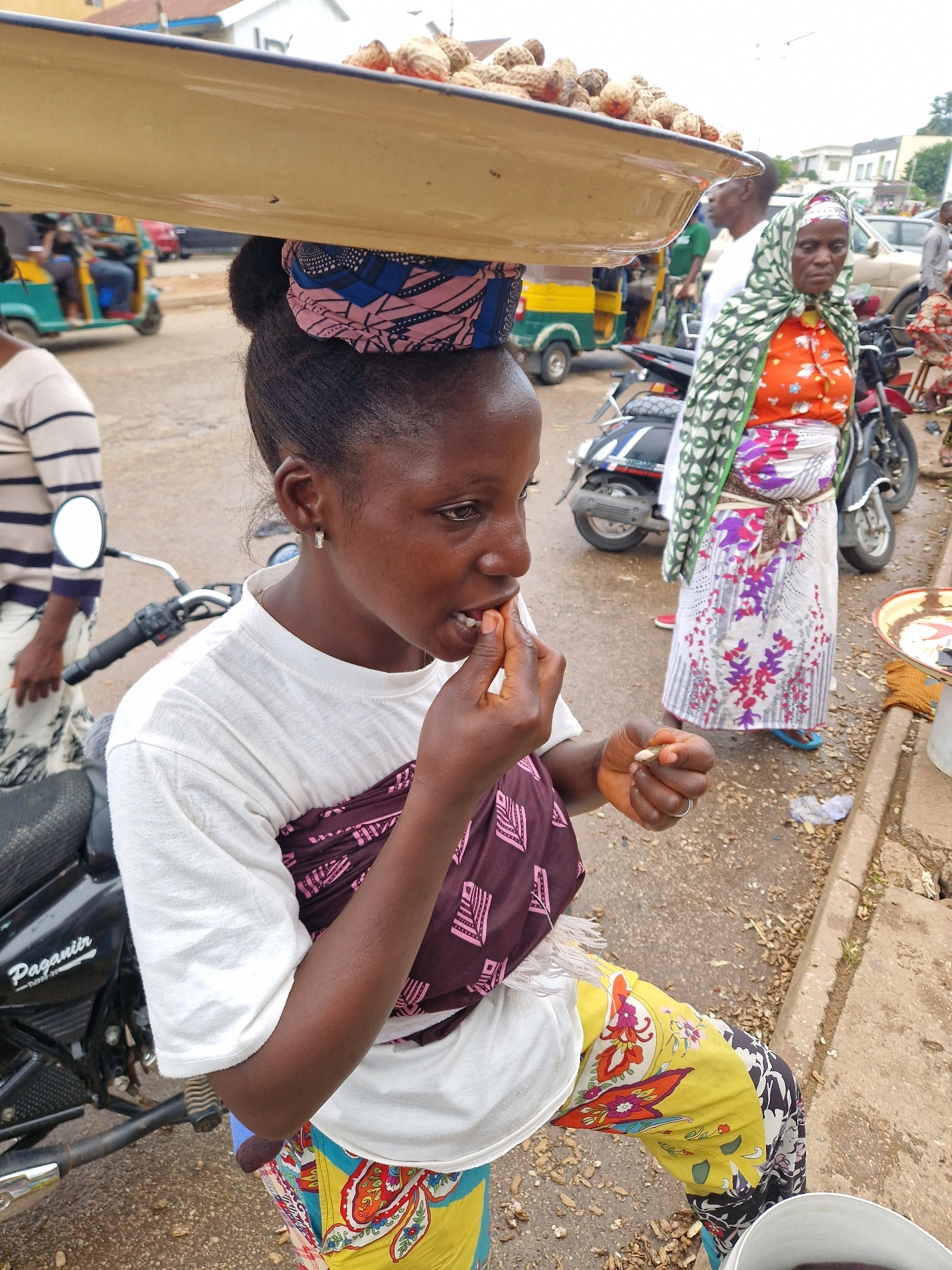 In Jos, Nigeria, a young woman balances a large metal tray filled with nuts on her head. She wears a white t-shirt with a patterned wrap around her torso and brightly colored, patterned trousers. She is actively eating a nut from her hand, bringing it to her mouth. In the background, a woman in a headscarf and patterned clothing stands near a motorcycle. Other vehicles, including a tricycle, are visible, suggesting a busy street scene. The ground is paved and appears wet, indicating recent rain. The overall environment suggests an outdoor market or roadside commerce.