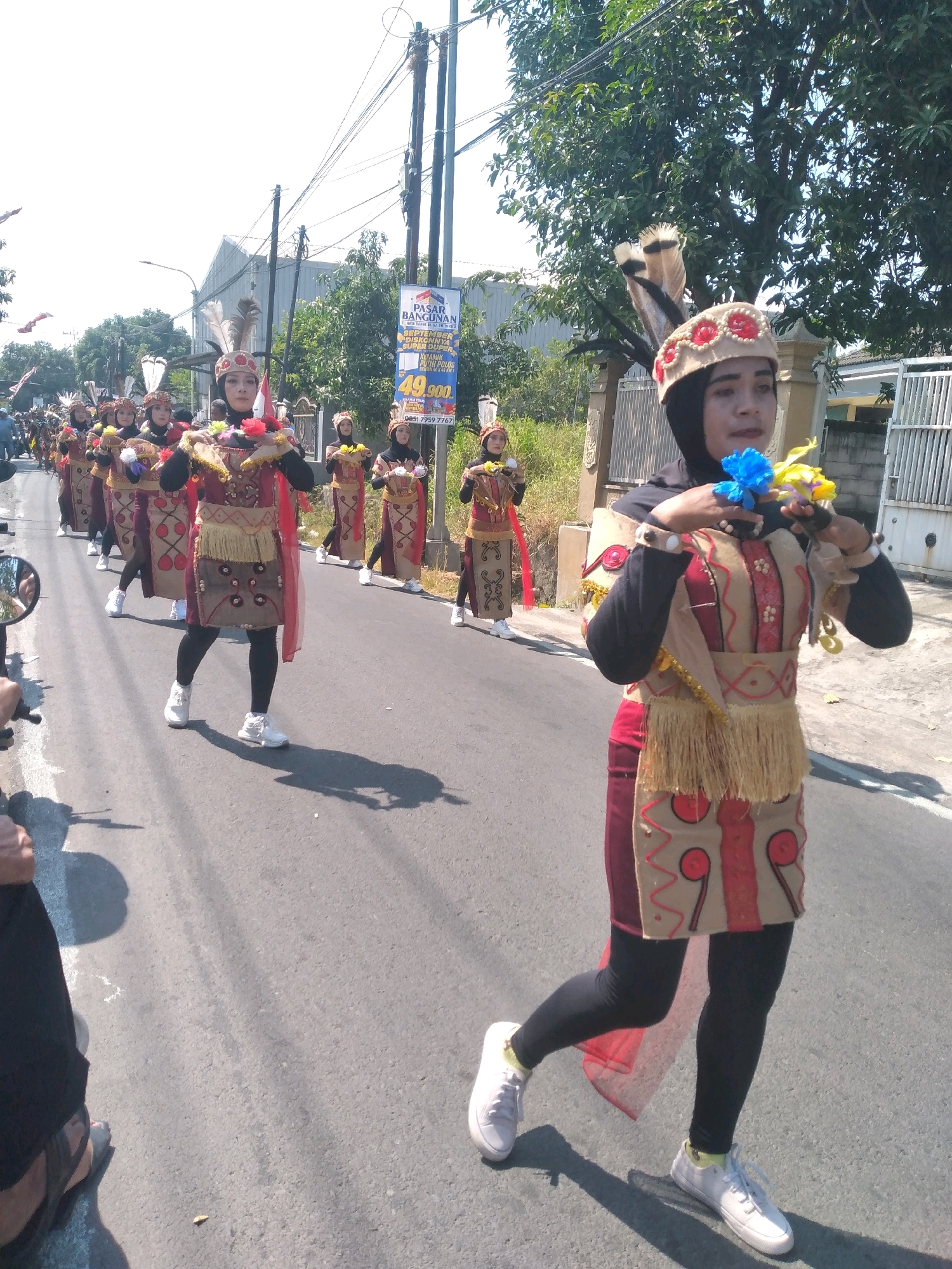 A group of women in traditional costumes are walking in a parade on a sunny day in Randegansari, Indonesia. The woman in the foreground is wearing a black hijab, a red and beige costume adorned with intricate patterns, and a headdress with feathers. She is holding a blue and yellow prop in her hands. Behind her, other women are also dressed in similar attire, forming a line. The parade appears to be taking place on a paved road with some buildings and trees visible in the background. A sign for "Pasar Bangunan" is visible on the right side of the image, indicating a building market. The overall scene suggests a cultural event or festival.