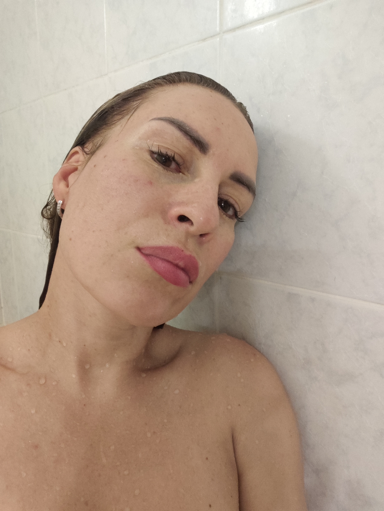 The image is a close-up, low-angle, self-taken photograph of a woman's face and upper torso, leaning against a white tiled wall. The woman has wet hair, suggesting she is in a shower. She is wearing a silver hoop earring. Her skin is speckled with water droplets. Her lips are painted with pink lipstick. The background is a wall of rectangular white tiles with visible grout lines. The image does not provide any specific visual cues to indicate a location in Almaty, Kazakhstan.