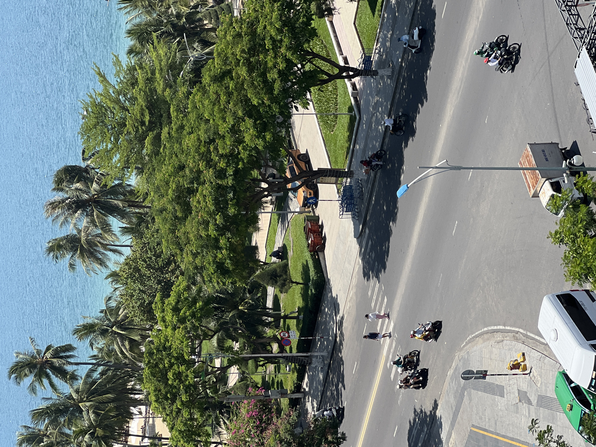 The image depicts a street scene in Nha Trang, Vietnam, with the turquoise waters of the ocean visible in the background. Palm trees line the coast, and lush green trees and manicured park areas flank the street. The street is busy with motorcycle traffic, and several people are crossing a crosswalk. A white van and a green taxi are parked on the side of the road, while a small truck drives in the distance. A street lamp and a fire hydrant are also visible. The sky is clear and blue, suggesting a sunny day.