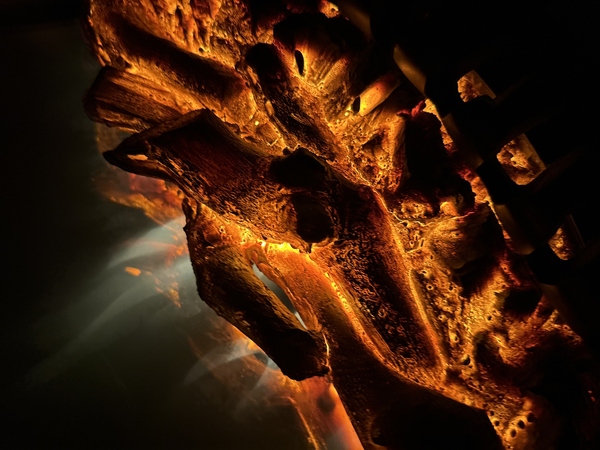 This is a close-up, low-angle shot of glowing embers and burning logs in a fireplace. The lighting is dramatic, with intense orange and yellow hues dominating the scene, creating a warm and fiery atmosphere. Wisps of smoke or steam are visible in the upper left portion of the image, adding a dynamic element to the static subject matter. The texture of the wood and ash is highly detailed, with rough surfaces and small pores visible. The background is dark and out of focus, further emphasizing the glowing elements. The overall impression is one of heat and intensity, capturing the raw power of fire. The image does not provide any information about the location in the city of Unknown, Unknown.