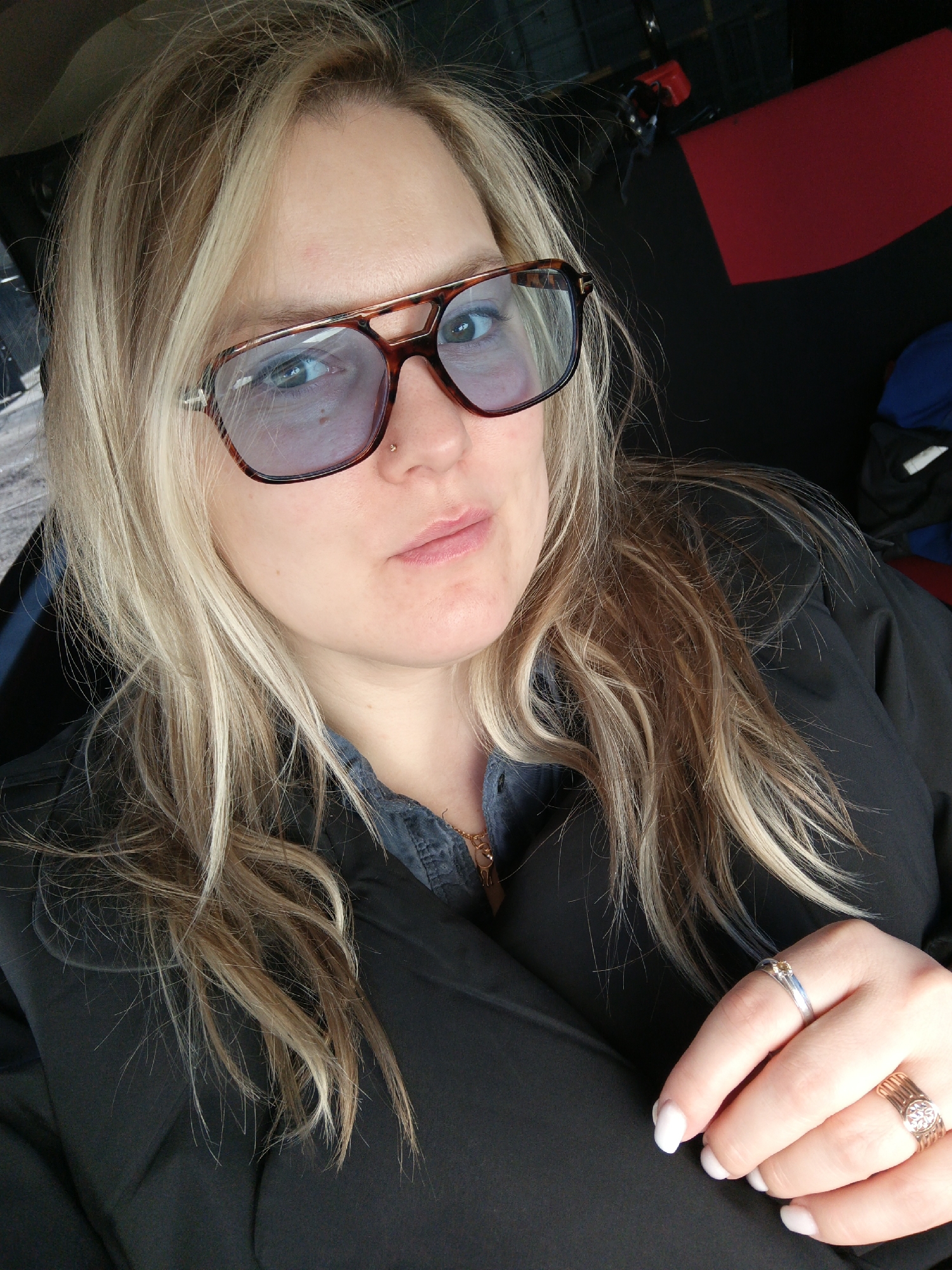 A medium shot shows a blonde woman with sunglasses. She is wearing a black jacket, a denim shirt, and jewelry. The location is difficult to determine definitively as Chaikovsky, Russia, based solely on the image. The background shows a car interior.
