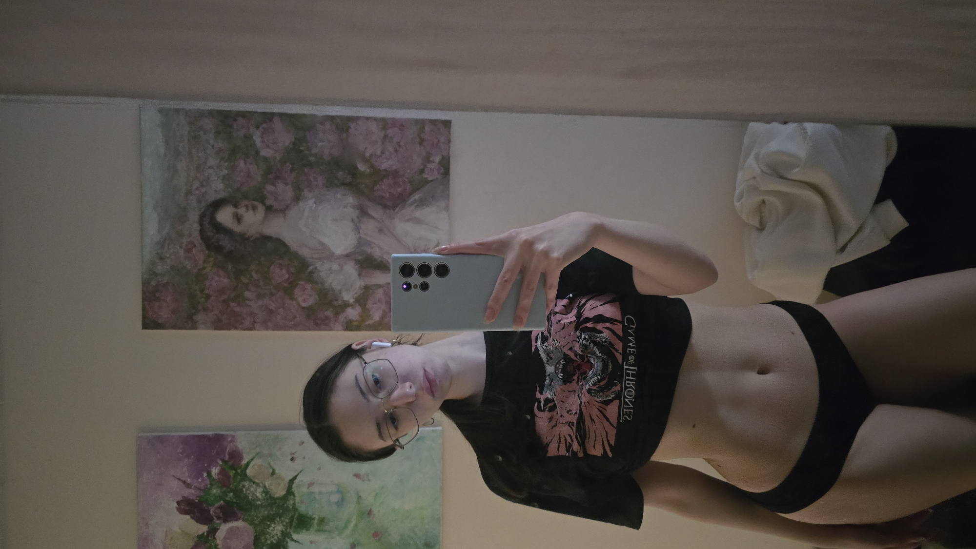 A woman takes a selfie in front of a wall adorned with paintings. She is wearing glasses, a black crop top, and black underwear. The paintings depict a woman lying in flowers and a bouquet of flowers. There is a pile of white and black fabric to the right of the frame. The specific location in Stara Zagora, Bulgaria is indeterminable from the image.