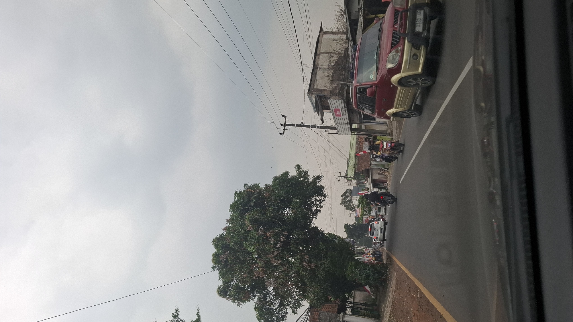 The image captures a street scene in Cilaku, Indonesia, under an overcast sky. The perspective is from inside a vehicle, looking forward through the windshield, which shows reflections of the interior. The road is paved with asphalt and has a yellow line marking the edge on the left and a white line in the middle.
On the left side of the road, there is a large, leafy tree with its branches extending over the road. Adjacent to the tree is a low brick wall and a building with a tiled roof. Several Indonesian flags are visible, mounted on poles along the roadside.
Traffic is present, with several vehicles and motorcycles. In the foreground on the right, a red and gold SUV is driving on the road. Further down the road, a white car is visible, and several motorcycles are being ridden by individuals wearing helmets. One motorcycle is being ridden by a person wearing a yellow shirt, and another by a person in a black shirt.
On the right side of the road, there are buildings with weathered facades and tiled roofs. Power lines crisscross the sky, supported by utility poles. The overall impression is of a typical urban street in Indonesia with moderate traffic and a somewhat cloudy atmosphere.