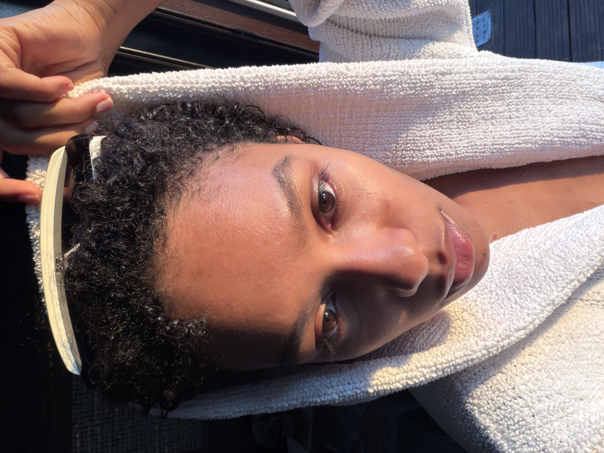 A close-up, eye-level, medium shot shows a young person with dark, curly hair and medium-toned skin, wearing a white terrycloth robe. The person is looking down and slightly to the left, with their head angled upwards, partially obscured by the hood of the robe. A comb is being held by their right hand, positioned on top of their head, with a few strands of hair visible between the comb and the scalp. The lighting suggests late afternoon or early morning sun, casting warm highlights on the person's face and the robe. The background is dark and out of focus, with a hint of wooden planks visible on the right side, suggesting an outdoor setting possibly on a deck or patio. There are no visible signs of the city of Champs-sur-Marne, France.