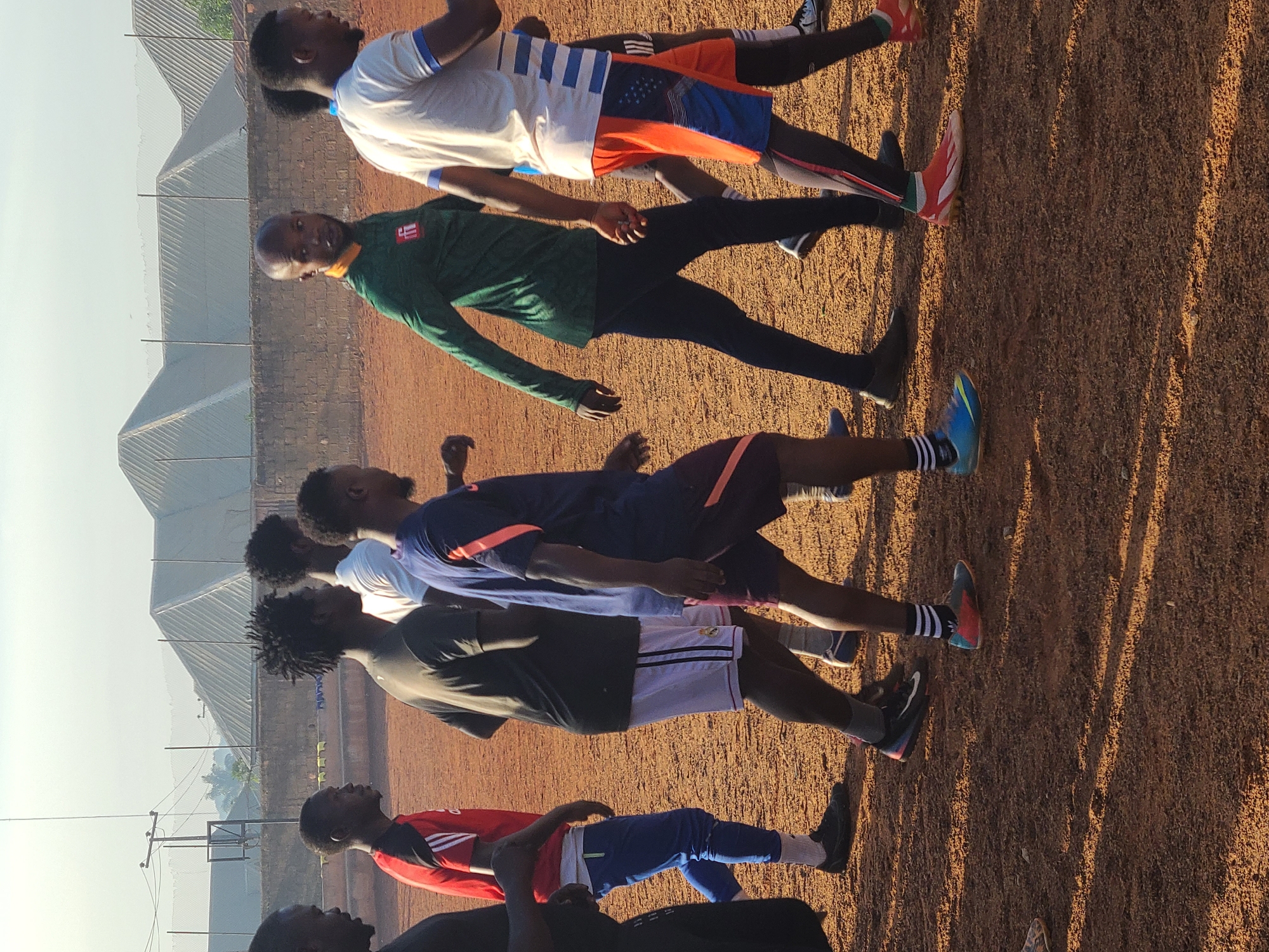 This media file captures a group of approximately 8-10 young adult males on a reddish-brown, sandy or dirt sports ground under clear skies. The scene is illuminated by warm, directional light, casting long shadows, indicating either early morning or late afternoon.
The subjects are dressed in various athletic wear, including t-shirts, shorts, track pants, and a variety of athletic shoes, suggesting they are involved in or preparing for a sports activity, likely soccer training. They are standing in various postures; some appear to be in a loose line, while others are in small clusters, possibly listening to instructions or waiting for the next part of their training session.
Notable details include:
- A man in a green long-sleeved shirt and black track pants is prominently featured, looking towards the viewer. His shirt bears a red and white emblem on the left chest, resembling a national or club flag/logo.
- Other men wear white, blue, black, and red athletic tops and various colored shorts or pants.
- Several pairs of athletic shoes are visible, including one with black and white striped socks featuring what appears to be an Adidas logo, and another pair of brightly colored cleats.
- In the background, there are low-lying buildings with corrugated metal roofs, a tall wire mesh fence, and distant hazy landscape features, common to an outdoor training facility in a semi-urban or rural setting.
The image depicts a moment of gathering or preparation during a sports session, likely in Jos, Nigeria, given the provided context.