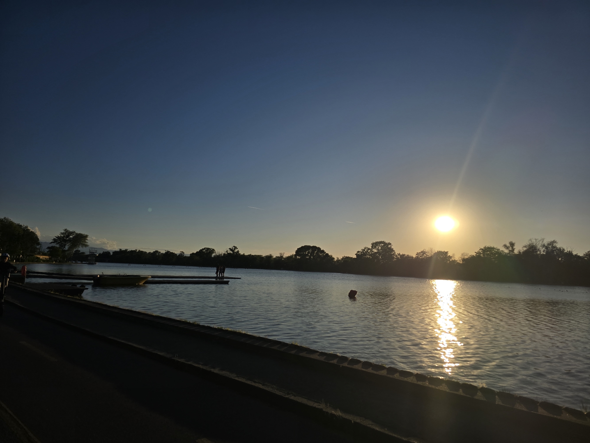 A landscape of a sunlit body of water, likely a lake, taken near a walkway in the city of Unknown, Unknown. The sun is shining, creating a bright spot on the water. Silhouettes of trees line the far shore. A small pier extends into the water with two people on it. Several boats are docked. There is one life preserver device visible in the water.
