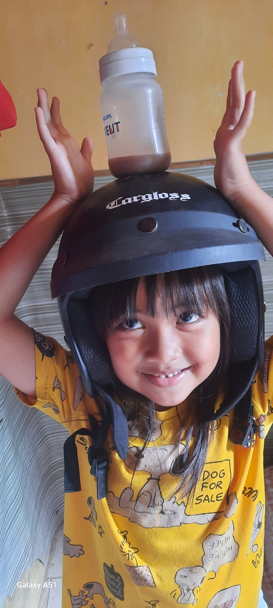 A young girl in Ploso, Indonesia, is balancing a baby bottle on a black helmet on her head. She is wearing a yellow t-shirt with a cartoon dog print. Her hands are raised above the helmet, palms facing outward, as if to keep it balanced. The baby bottle is white with a brown liquid inside. The background is a yellow wall. The image appears to be taken indoors during the daytime. The girl has a cheerful expression on her face. Visible text includes "Philips AVENT" on the bottle, "largloss" on the helmet, "DOG FOR SALE" on her t-shirt, and "Galaxy A51" in the bottom left corner.