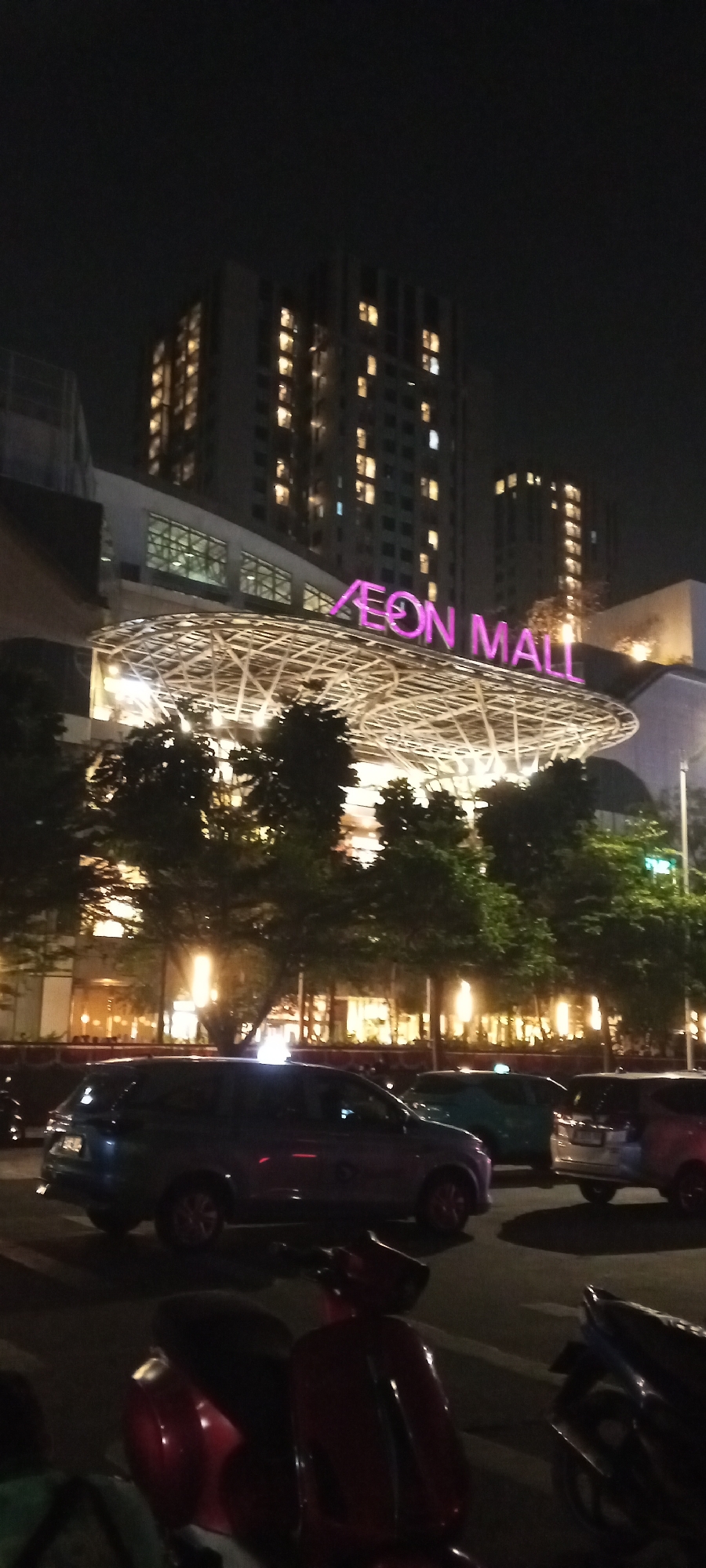 This is a nighttime photograph taken in South Jakarta, Indonesia, showing the AEON Mall. The mall features a large, illuminated sign with pink lettering that reads "AEON MALL" above a modern, white, arched structure. Behind the mall, several tall buildings are visible, with many windows lit up, suggesting they are residential or office spaces. In the foreground, there are parked cars and a red scooter. The overall impression is of a bustling urban commercial center at night.