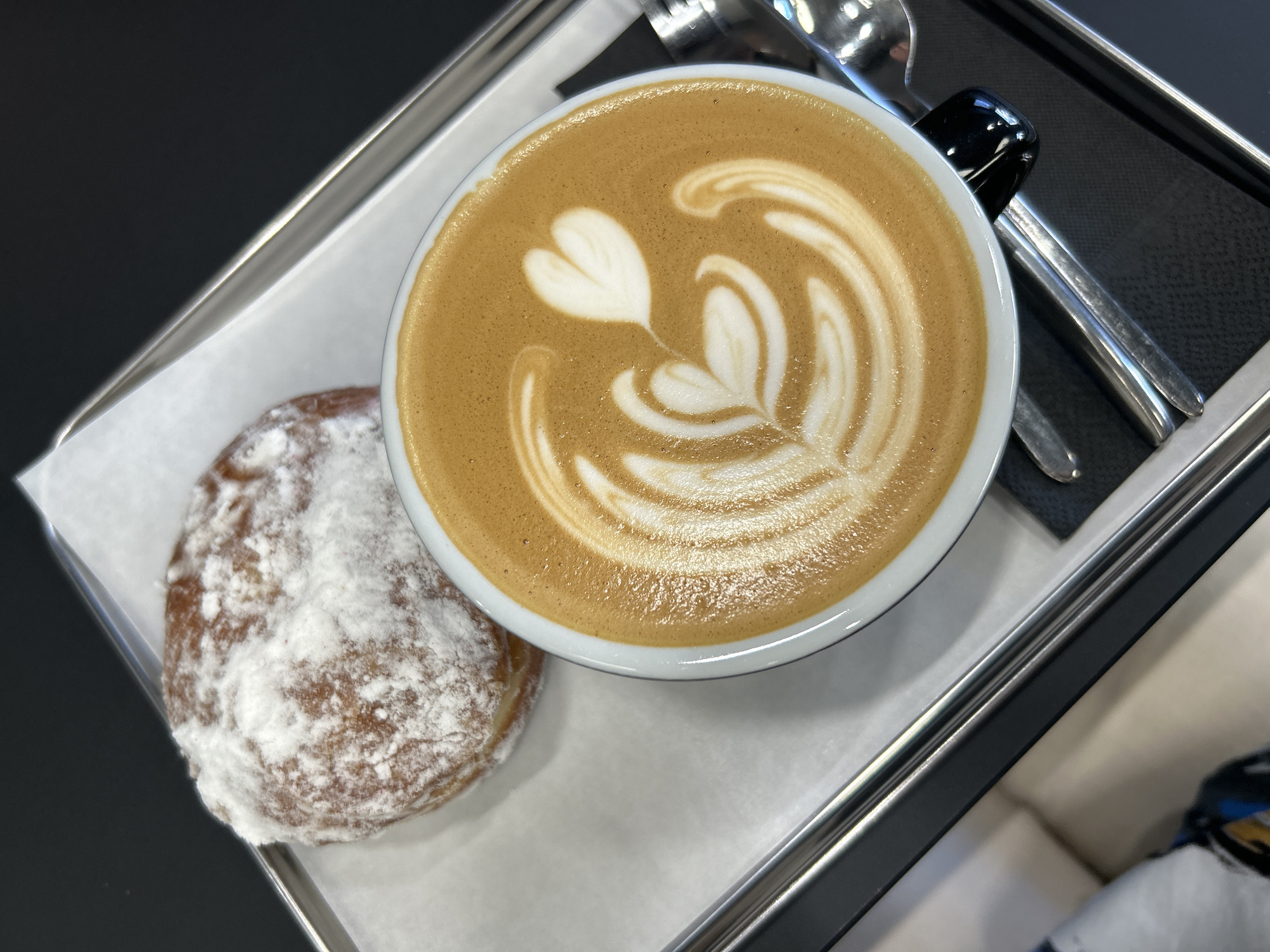 The image shows a coffee and a donut on a metal tray. The coffee is in a white cup with a black handle and features latte art with a heart and leaf design. The donut is round and covered in powdered sugar. A spoon rests alongside the cup on the tray, which is lined with white paper. The background is out of focus, but suggests a modern interior setting. The location, Izhevsk, Russia, cannot be verified from the image.