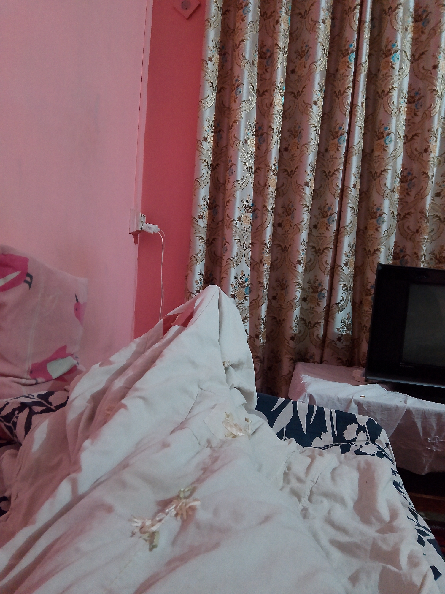 The image shows a messy bed in a room with pink walls. A white duvet is rumpled and draped over the bed, with a pink pillow visible on the left. A floral curtain hangs in the background, and a television set is placed on a table to the right. An electrical outlet with a cord plugged into it is mounted on the wall. The scene appears to be within a residential setting in Mansehra, Pakistan.