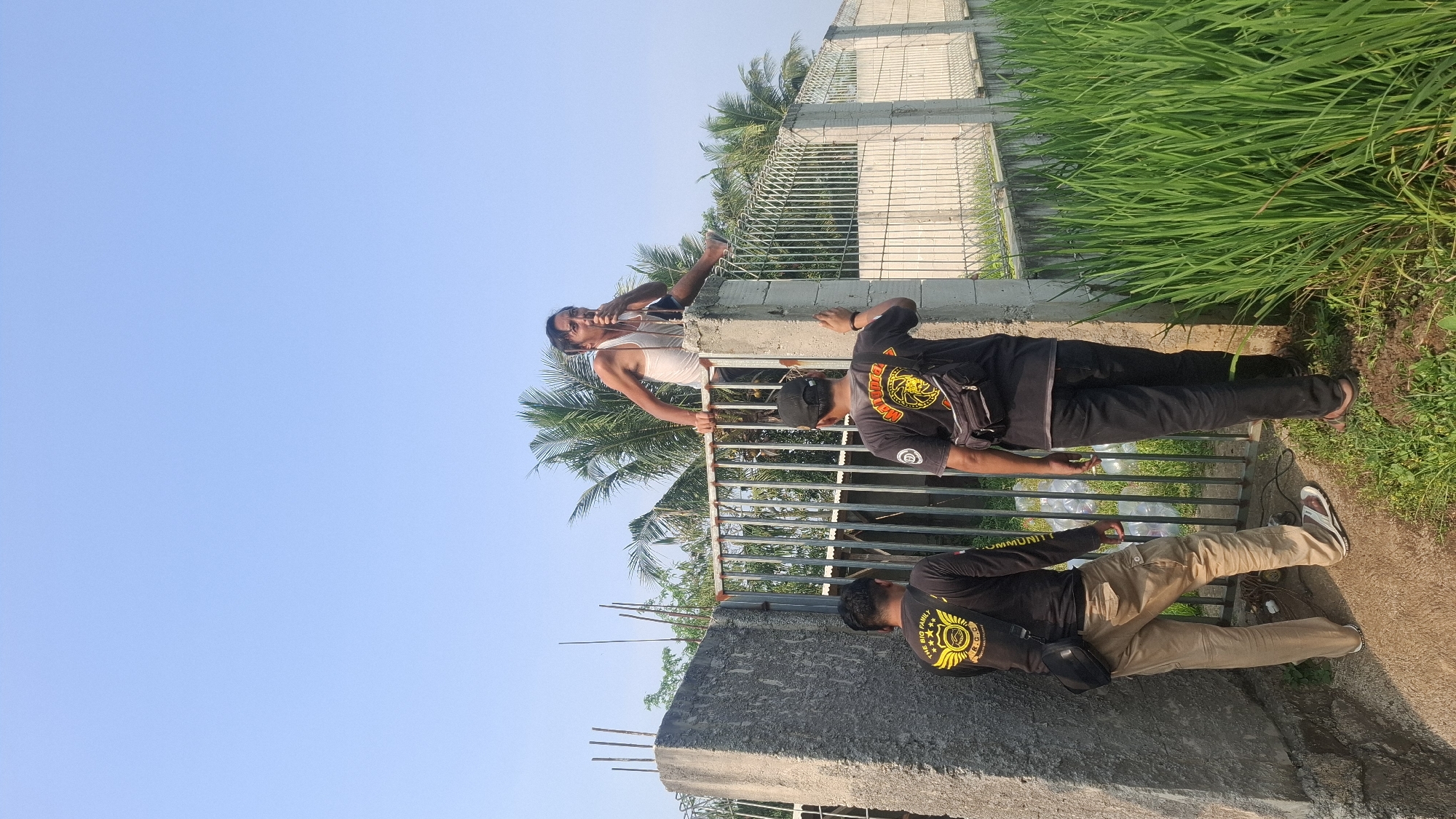 In Rahong, Indonesia, on a bright, clear day, three men are present. One man, wearing a white tank top, is perched atop a concrete fence post, holding a cigarette to his lips and speaking into a microphone. He appears to be leaning on the metal gate. Below him, two other men stand facing away from the camera, their backs turned towards the gate and the man on the post. Both are wearing black t-shirts with yellow emblems. The man on the left wears tan cargo pants and sandals, while the man on the right wears black pants and sandals. A lush green field of tall grass borders the right side of the image, with more greenery and palm trees visible in the background. The concrete structure on the left is likely part of a building or property boundary.
