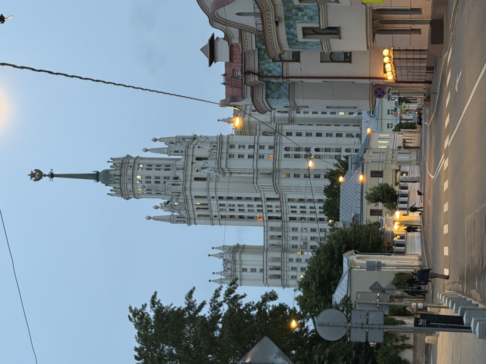 The image captures a street scene in Moscow, Russia, featuring the Kotelnicheskaya Embankment Building, one of the "Seven Sisters" skyscrapers built in the Stalinist architectural style. The building's ornate facade, characterized by its white stone and spires, dominates the left side of the frame. A wire runs across the top left, and a street lamp adds illumination.
On the right is a street with crosswalk lines and a building with colorful tiled details and lighted orbs. A crosswalk with multiple pedestrians is present with more people on the right sidewalk. Street signs and trees are in the foreground. The sky is dusky, suggesting the time is either dusk or dawn. The setting is urban, with a mix of architectural styles visible.