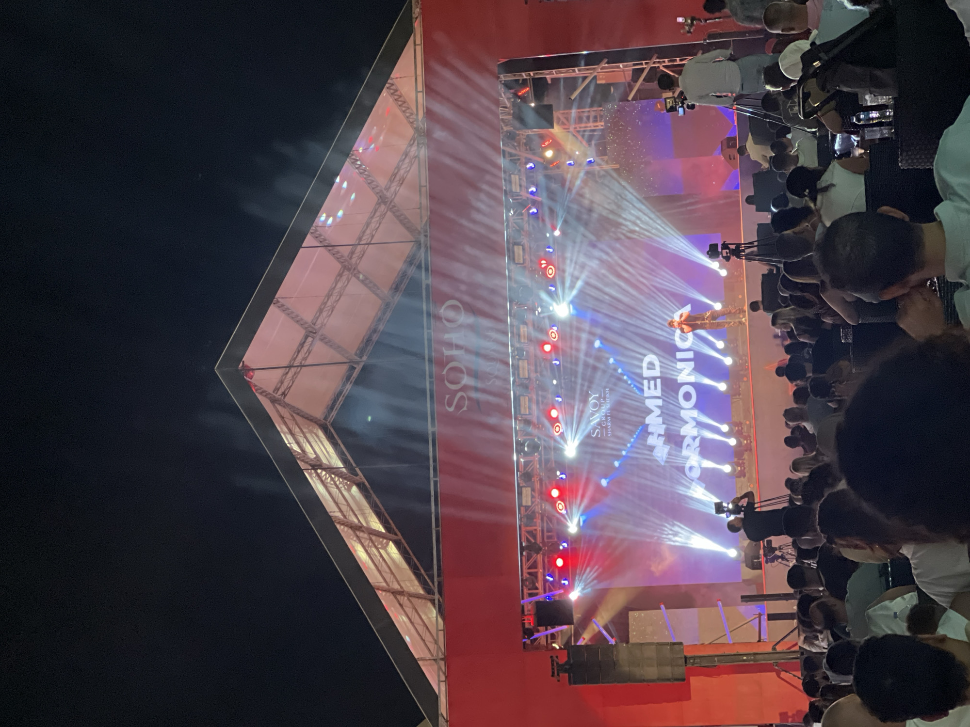 An outdoor nighttime concert is taking place at the Savoy Group in Sharm El Sheikh, Egypt. A large stage is illuminated by bright spotlights and is adorned with the text "SOHO" and "SAVOY GROUP - SHARM EL SHEIKH" along with "AHMED ORMONICA". A performer, identified as Ahmed Ormonica, stands on stage. Several cameras and camera operators are present, filming the performance. A large audience is seated in front of the stage, facing the performers. The environment appears to be a resort or entertainment venue with a large, open-air setup.