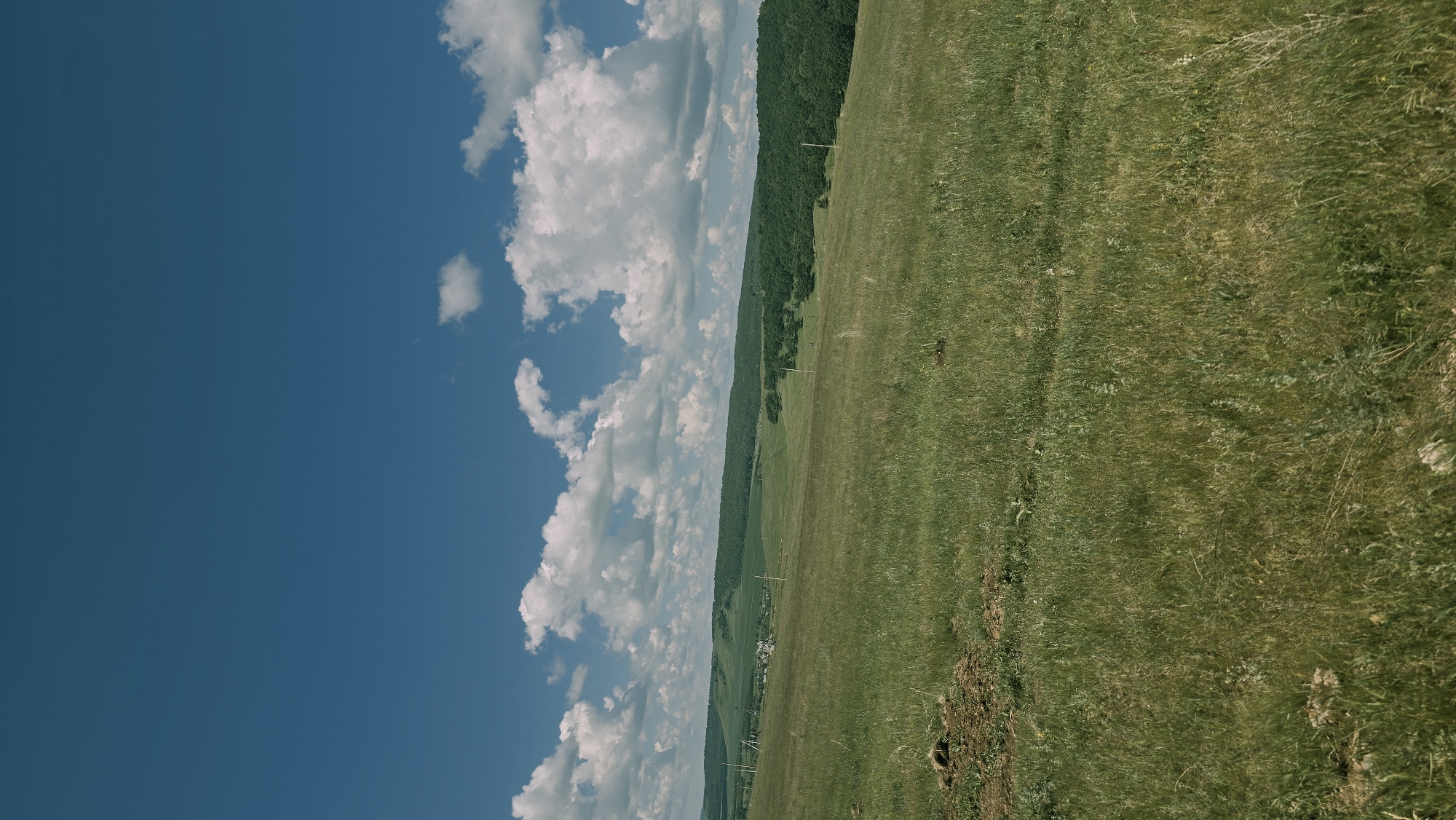 The image shows a panoramic view of a hilly landscape under a bright blue sky with scattered white clouds. The foreground consists of a grassy field, with some patches of dry earth visible. In the mid-ground, there are rolling green hills dotted with trees and a small cluster of buildings, suggesting a rural settlement. Several utility poles with wires traverse the landscape. The background is dominated by more hills covered in dense green forests. The scene appears to be in Kudashevo, Russia, a village known for its natural beauty and agricultural land. The lighting suggests it is daytime, possibly late morning or early afternoon, with the sun casting soft shadows. There are no people or animals visible in the image.