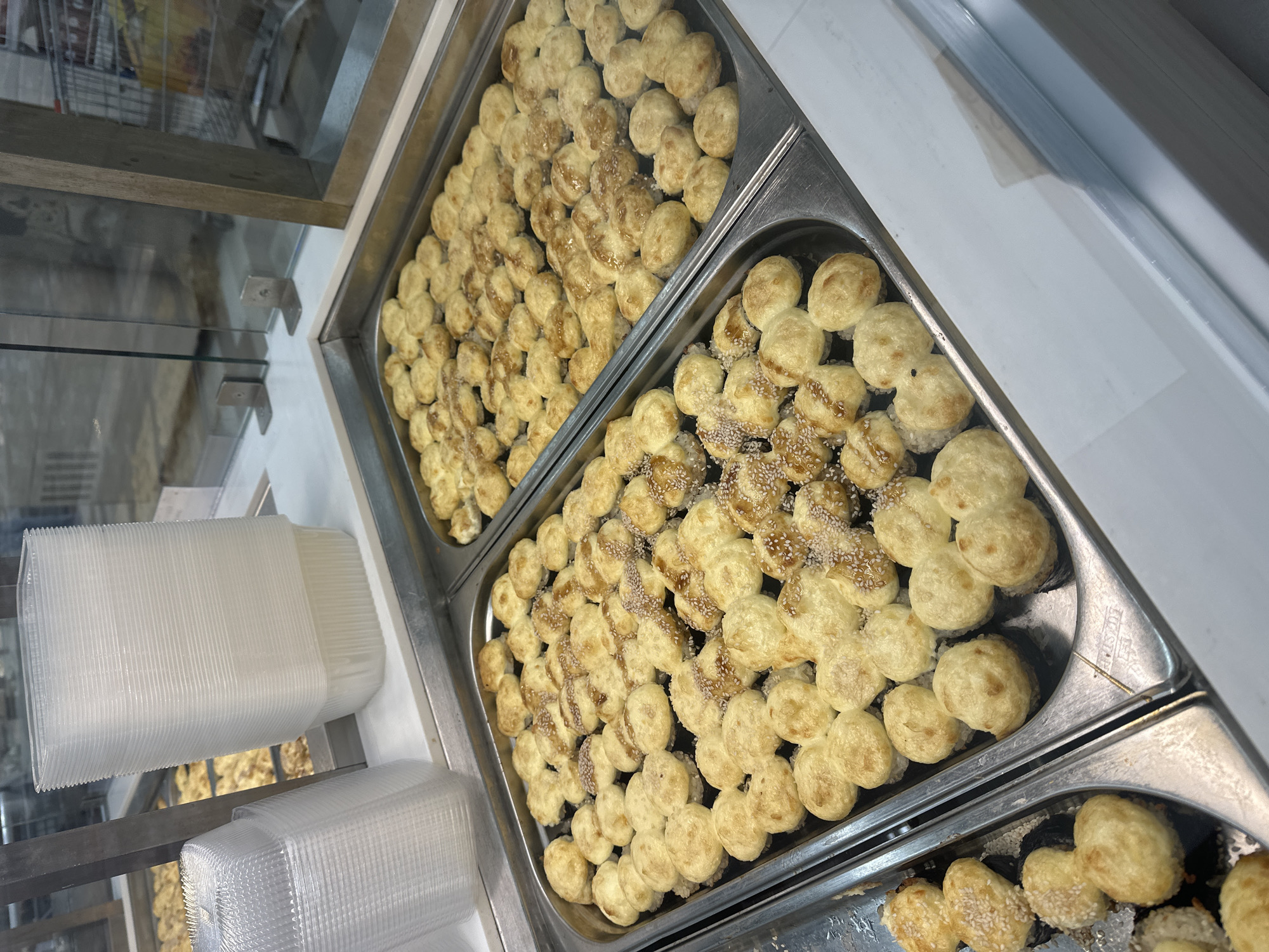 The image shows two stainless steel trays filled with what appear to be baked goods, possibly small buns or pastries, some sprinkled with seeds (likely sesame) and possibly a light glaze. The trays are in a glass display case, likely in a bakery or food service establishment. To the left of the trays, partially visible, are stacks of clear plastic containers. The items are arranged neatly in rows, suggesting a commercial setting. The image does not contain any people. There is no information in the image to suggest the location in the city of Nizhny Novgorod, Russia.