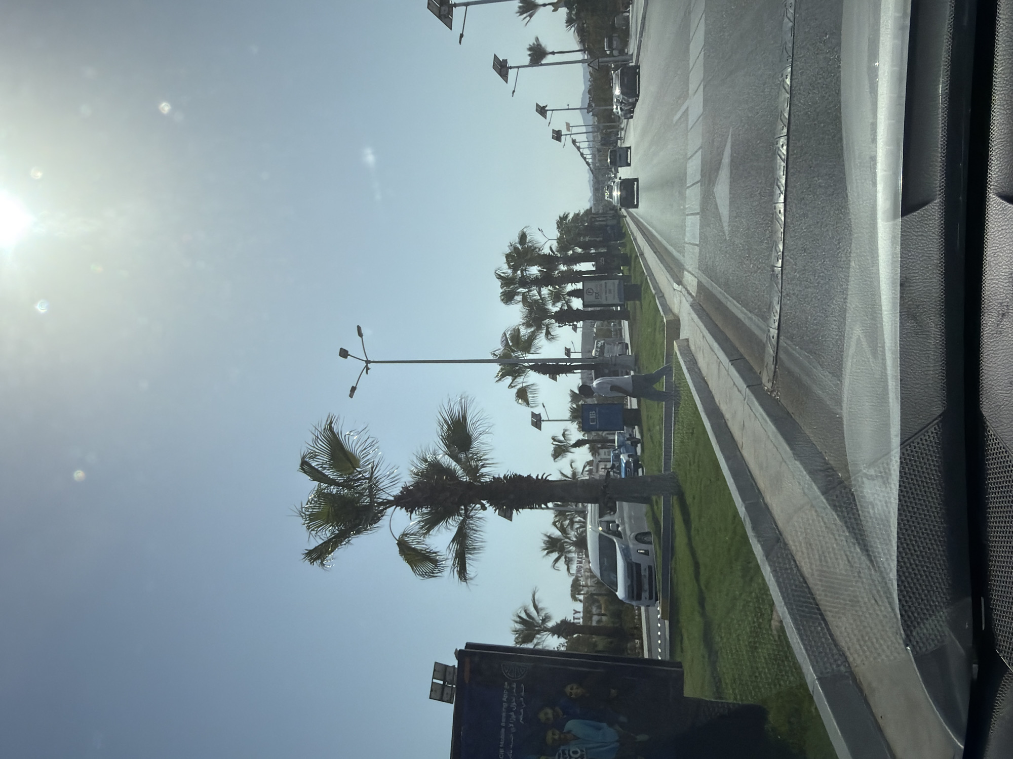 The image captures a sunlit street scene in Sharm El Sheikh, Egypt. The perspective appears to be from inside a vehicle, looking forward. Palm trees line the median and sides of a multi-lane road. Several vehicles, including a white van, a blue pickup truck, and cars, are visible on the road. Pedestrians are present on the sidewalk. One man in a light-colored shirt and pants walks on the sidewalk. A large billboard is visible on the left side of the frame. Streetlights with solar panels are spaced along the road. The sky is clear and bright, with the sun creating a lens flare effect. The environment suggests a warm, tropical climate, consistent with Sharm El Sheikh.