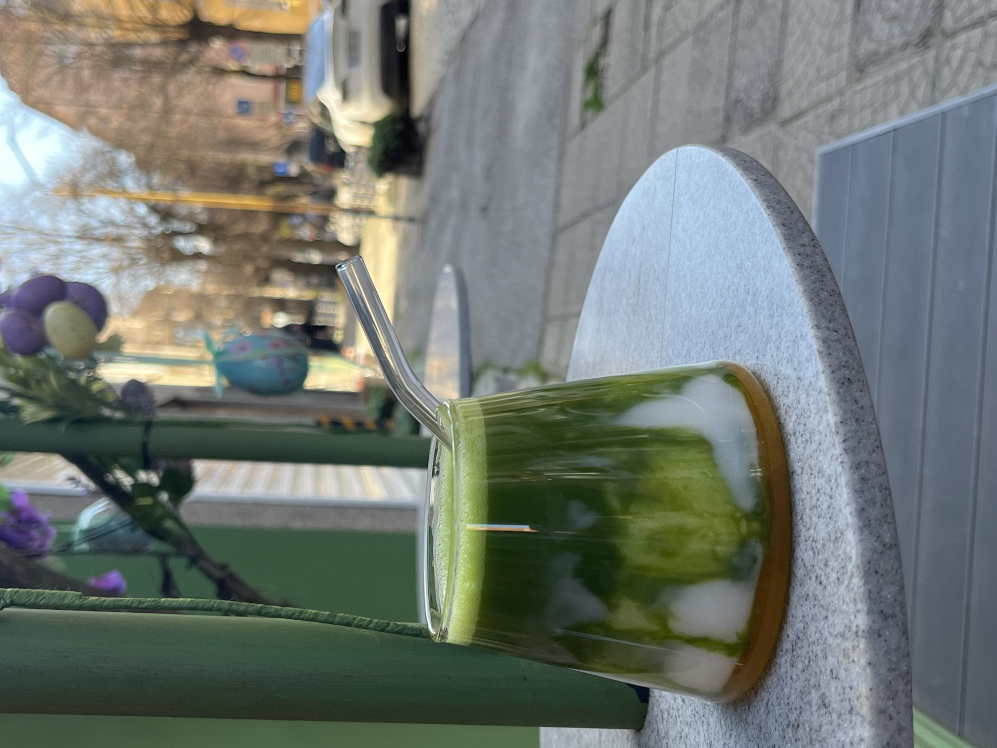 A close-up shot captures a glass of green liquid with a metal straw, likely a beverage like matcha latte, positioned on a round, speckled table. The table is placed outdoors, likely on a patio or sidewalk, given the partial view of an adjacent slatted gray table.
In the blurred background, an urban streetscape unfolds. There are buildings, trees, and a white vehicle parked on the street, suggesting a residential or mixed-use area. A decorative arrangement, possibly flowers or other ornaments, can be seen near the edge of the frame. The image seems to have been taken outdoors, and daylight is present.
The image does not provide definitive information that it was taken in Sofia, Bulgaria.