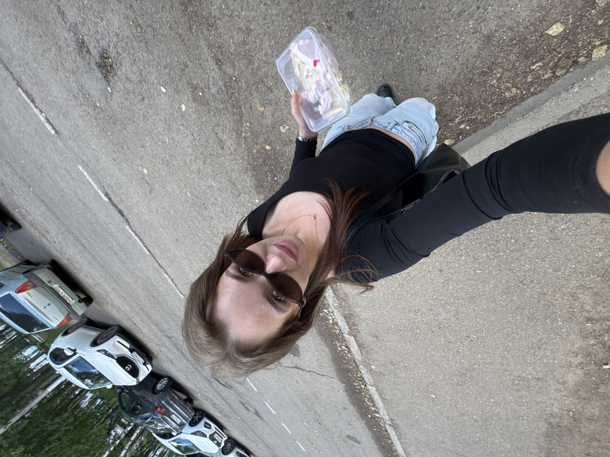A woman in sunglasses and a black long-sleeved top stands on a paved surface, holding a clear plastic container of food. She is wearing light blue jeans and black shoes. In the background, several cars are parked on the side of a road lined with trees. The scene appears to be an outdoor location in the city of Chaikovsky, Russia.