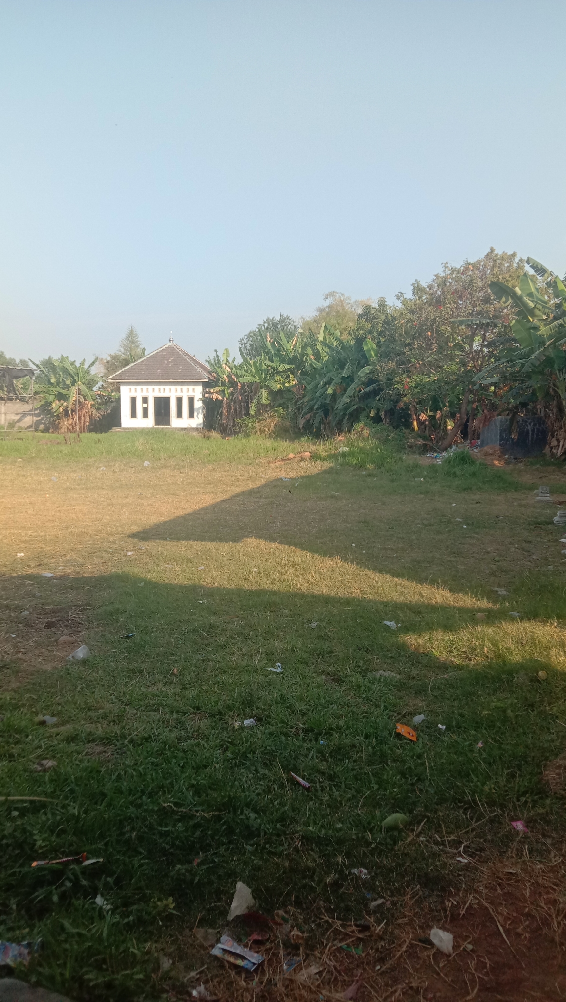 The image shows an outdoor scene in Karanganyar, Indonesia. In the mid-ground, a small, white building with a dark, tiered roof stands against a backdrop of lush green trees, including banana trees. The building has several windows and a prominent entrance. The foreground consists of a grassy field, partially dry and partially green, with some scattered litter visible on the ground. A long shadow stretches across the field from the right side of the frame, indicating it is daytime. The sky is clear and light blue.