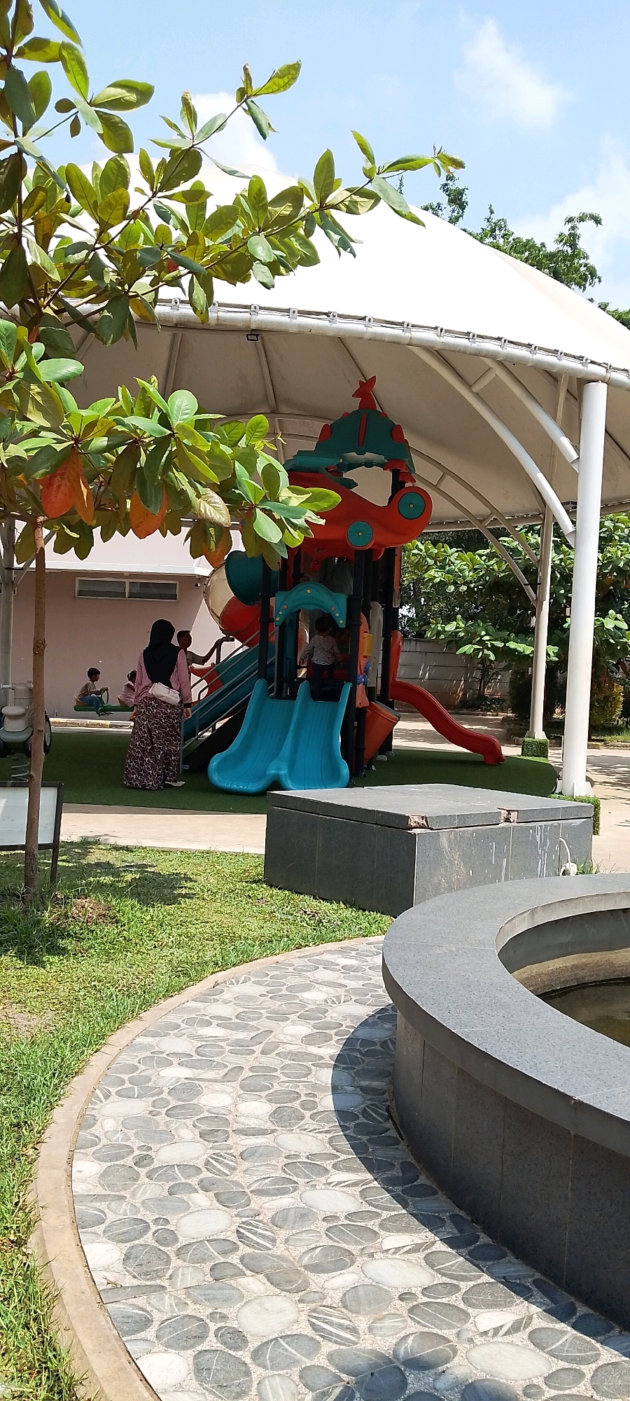 In the city of Sriamur, Indonesia, a brightly colored playground structure is situated under a large white canopy, providing shade. The playground features multiple slides, including two blue ones, a red slide, and various climbing elements. A child is visible playing on the blue slides. To the left of the playground, a woman wearing a hijab and a patterned skirt stands near a building. Two other children are seated on a seesaw-like toy on the grass in front of the building. The ground around the playground is covered with green artificial turf, and a winding pathway made of pebble-like stones leads towards the foreground of the image. In the foreground, a curved stone structure, possibly a fountain basin, is partially visible. Lush green trees and foliage surround the playground area, and the sky above is blue with scattered white clouds.