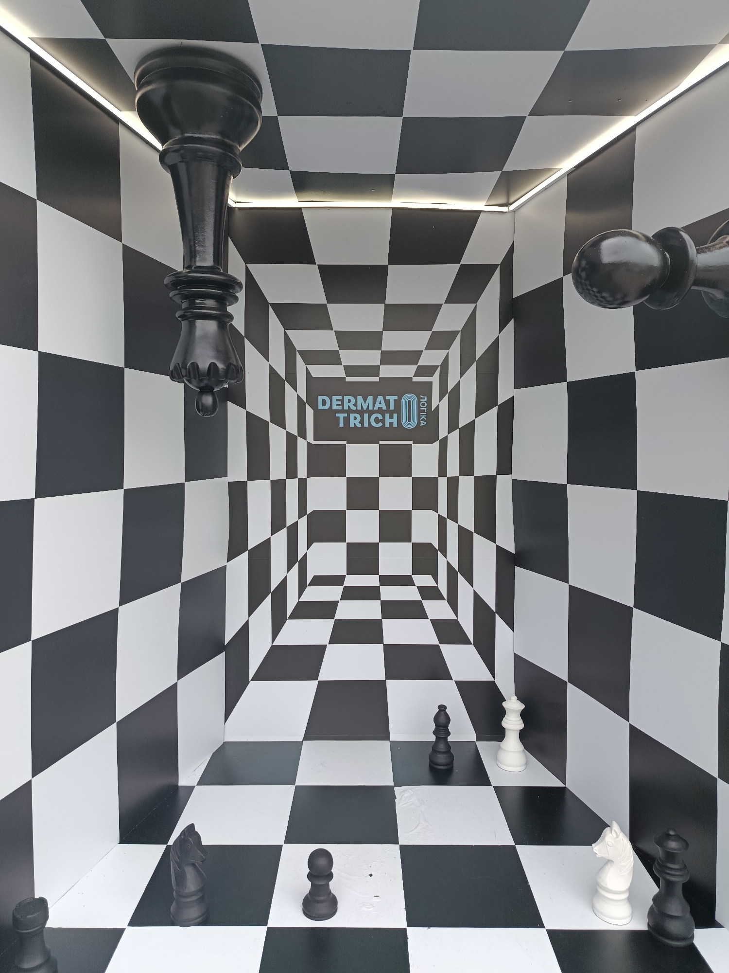 The image depicts an optical illusion room or art installation designed like a giant chessboard, located in Vynnyky, Ukraine. The scene utilizes forced perspective, with the black and white checkerboard pattern covering the floor, walls, and ceiling, converging towards a central point on the back wall, creating an exaggerated sense of depth.
Oversized chess pieces are strategically placed throughout the room. On the floor, closer to the viewer, are several black and white pieces: a black rook and a black pawn are visible on the bottom left, while a white knight and a black queen stand on the bottom right. Further into the room, a black knight and another black pawn are seen on the left side, and a white queen and a black bishop on the right. Adding to the surreal effect, large black chess pieces are affixed to the ceiling in an inverted position; a prominent black king hangs from the top-left corner, and part of another black piece, possibly a pawn or rook, is visible upside down on the top-right.
LED strip lighting runs along the seams where the ceiling meets the walls, illuminating the space and highlighting the architectural lines. On the distant back wall, centered in the converging checkerboard pattern, the light blue text "DERMATRICH" is displayed horizontally, with the word "ЛОГІКА" (Logika, meaning "logic" in Ukrainian) oriented vertically to its right. There are no people visible in the scene. The overall impression is one of an immersive, mind-bending environment playing with scale and perspective.