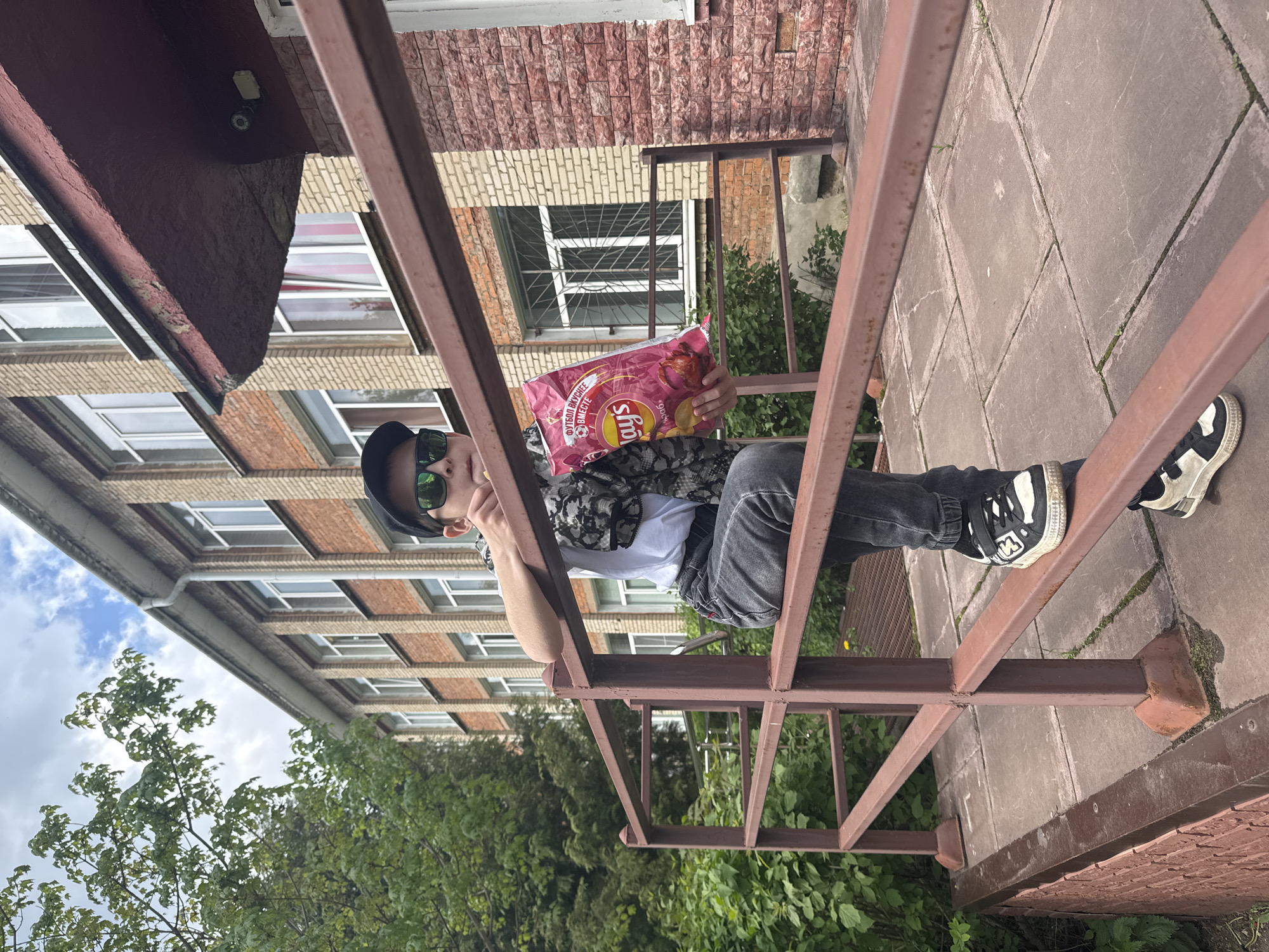 A person is sitting on a metal railing in Shevlyakovo, Russia, holding a bag of Lays potato chips. The person is wearing a white t-shirt, dark gray jeans, black and white sneakers, a black cap, and green sunglasses. Behind the person is a brick building with windows and greenery. The railing is brown. The bag of chips is predominantly red and features images of potatoes. The background includes trees and a partly cloudy sky. The image appears to be taken from a low angle.