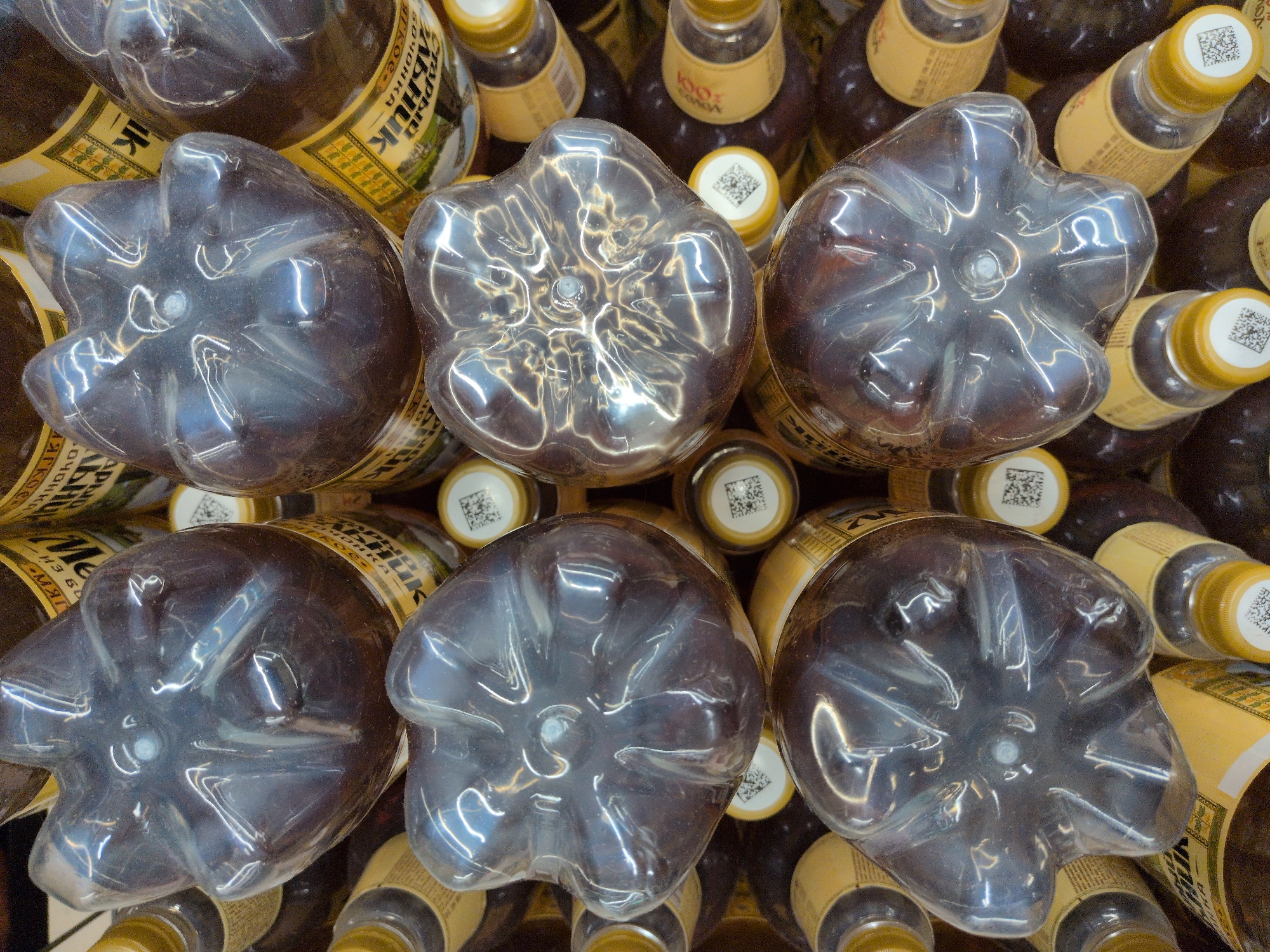 The image provides an overhead view of numerous dark plastic bottles, likely containing a beverage, stacked closely together. The foreground is dominated by the bottoms of several bottles, each featuring a distinct five-lobed or star-shaped indent design. Reflections of light shimmer across their glossy surfaces.
In the background and between the prominent bottle bases, the necks and yellow caps of other bottles are visible. Many of the yellow caps are adorned with a white circular sticker featuring a QR code. The labels wrapped around the bottles are predominantly yellow or beige and display Cyrillic text.
Visible text on the labels includes "МЕЛЬНИК" (Melnik), "БОЧОНКА" (bochonka, meaning 'small barrel' or 'keg'), and partial words like "ИК". One smaller label also shows "100% COK" (100% juice). Given the bottle design and the text "БОЧОНКА", these bottles are commonly associated with kvass or beer in Russia. The dark amber liquid inside further supports this.
The arrangement suggests the bottles are on a shelf in a store or a storage area. There are no people present, and the time of day or specific weather conditions are not discernible. The scene implies a commercial or retail setting in Sochi, Russia, consistent with the visible Cyrillic branding.