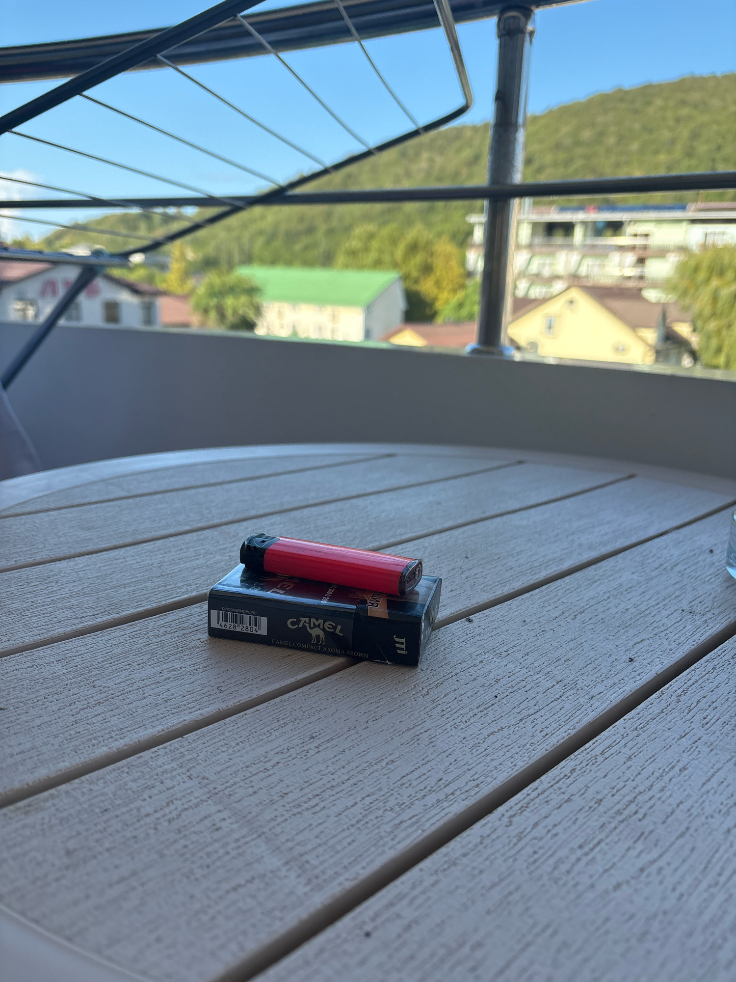 A red cigarette lighter rests on top of a pack of Camel cigarettes on a white wooden table outdoors. In the background, a lush green hillside with buildings and trees is visible under a clear blue sky. A railing and a clothes drying rack are also in the foreground. The scene appears to be on a balcony or patio in Arkhipo-Osipovka, Russia, a resort town on the Black Sea coast. The lighting suggests it is daytime.