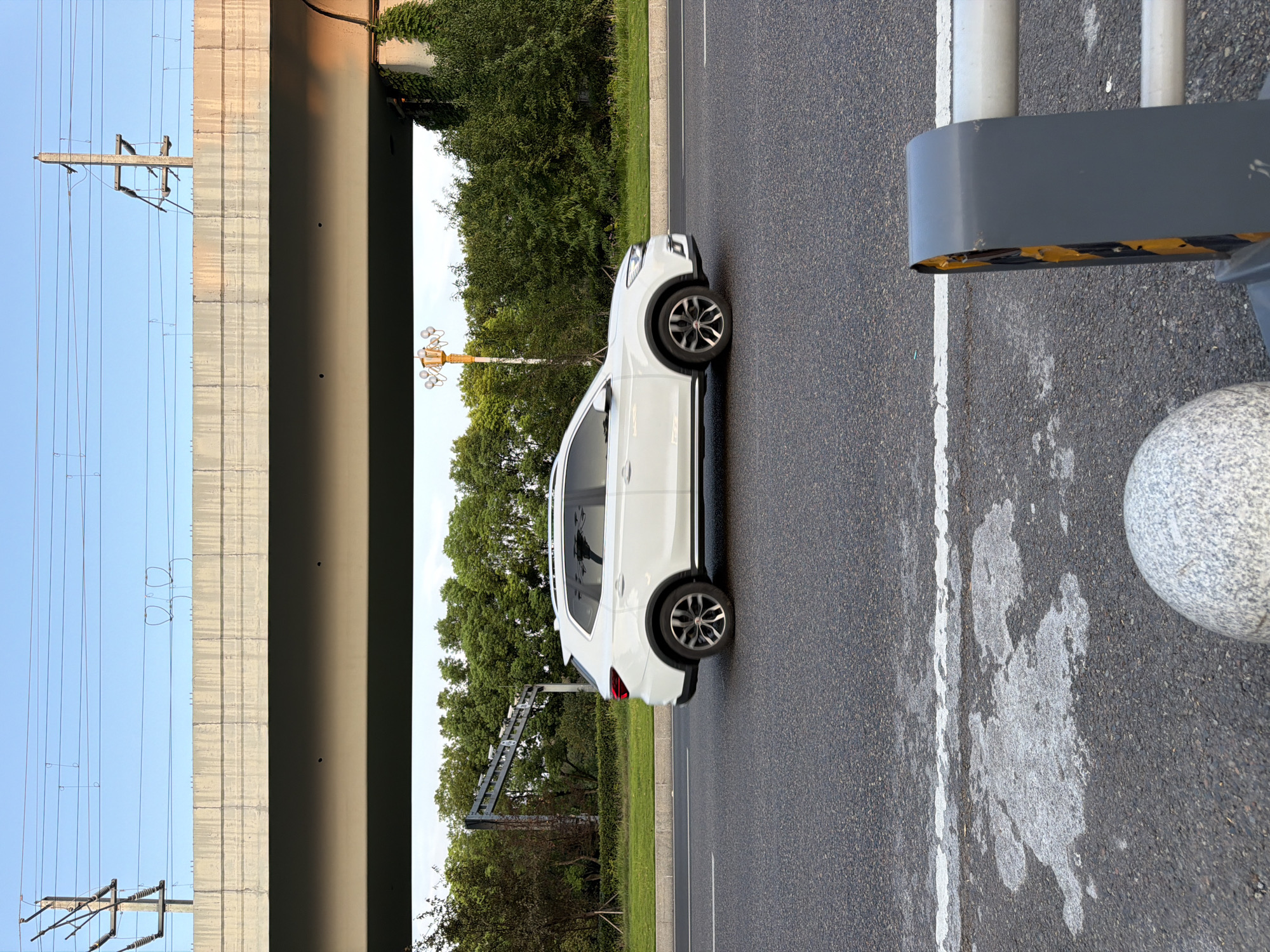 A white SUV drives on a street in Jiangjin District, China, under a highway overpass. The overpass has power lines running above it. Trees and greenery line the side of the road, and a streetlamp is visible in the distance. In the foreground, a concrete bollard and part of a barrier are visible. The sky is clear and blue.